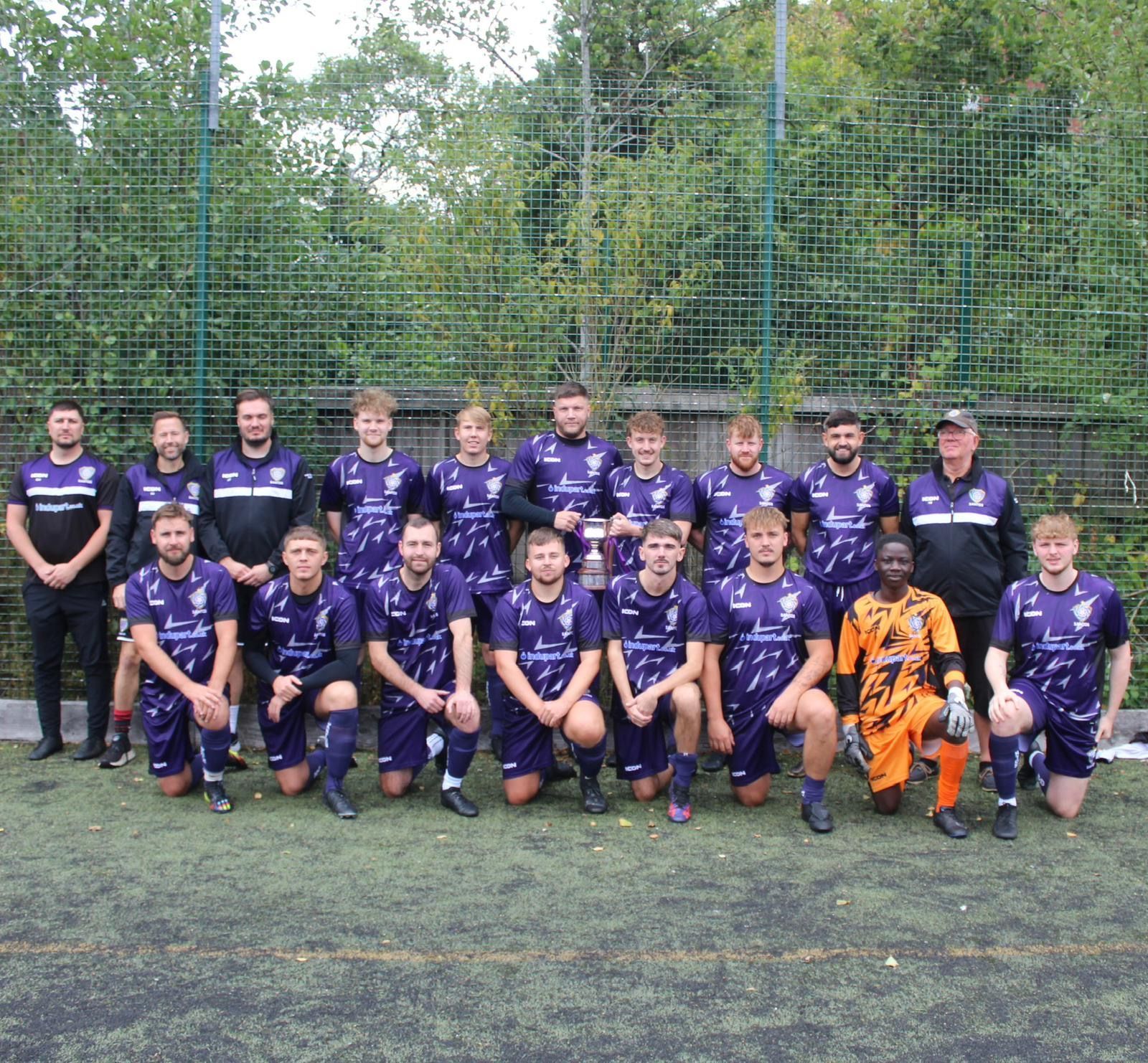 Youth soccer team posing on a field in purple uniforms, with one player in orange gear in front.