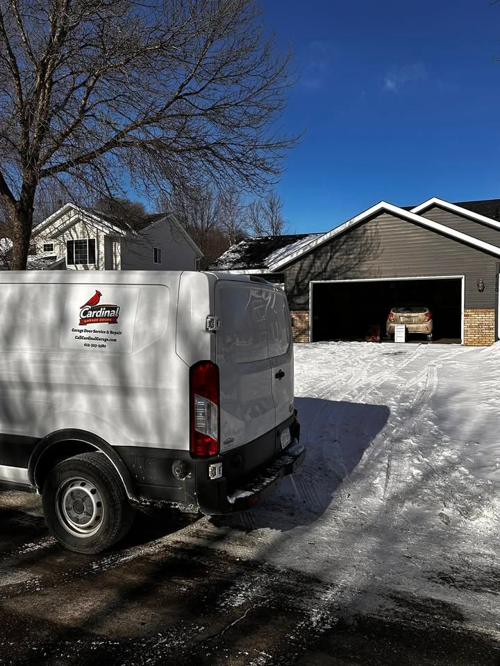 White utility van parked on a snowy driveway beside a house with an open garage under a clear blue sky