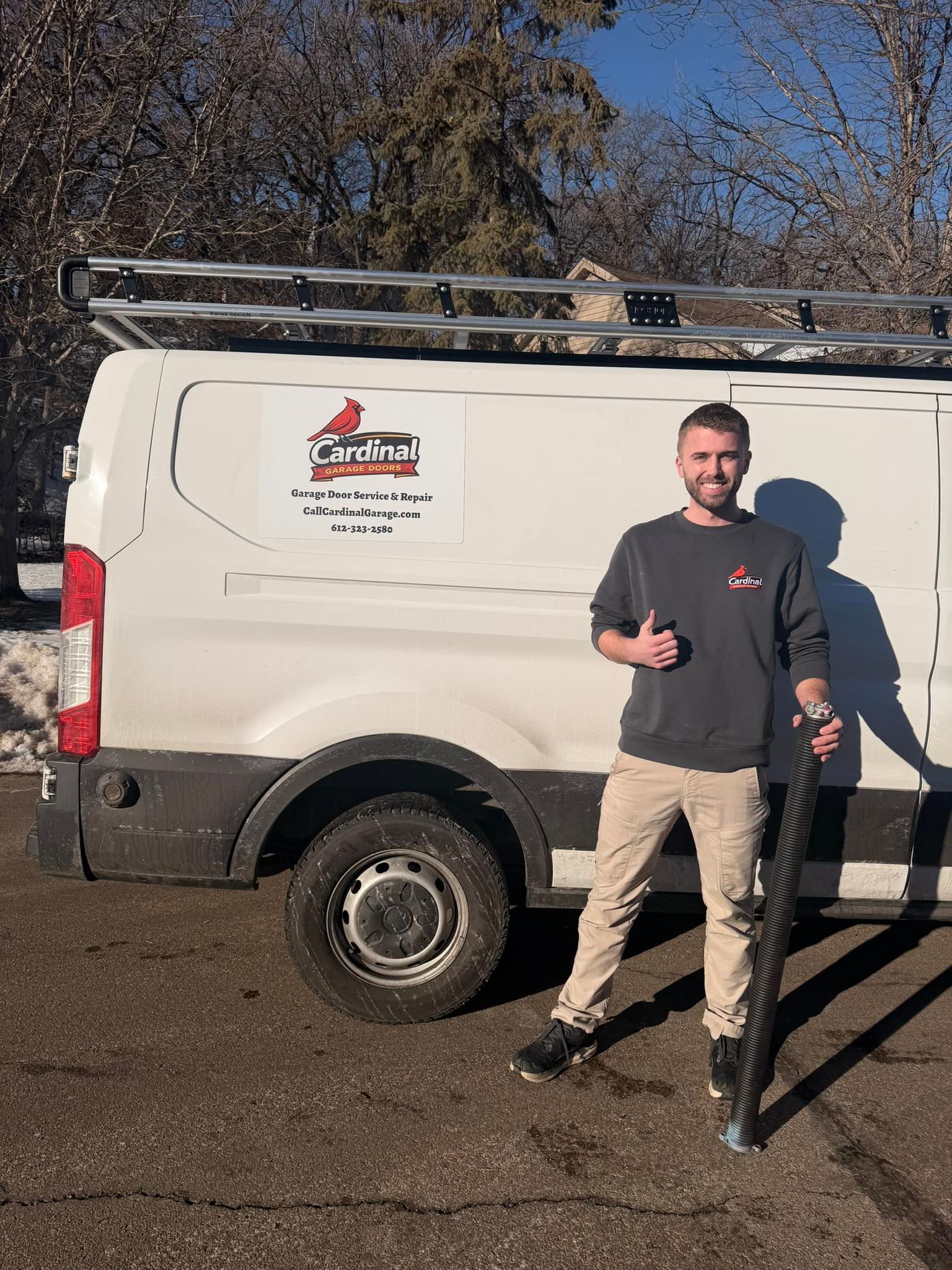 Man standing beside a white work van with a ladder rack in a sunny parking lot, giving two thumbs up