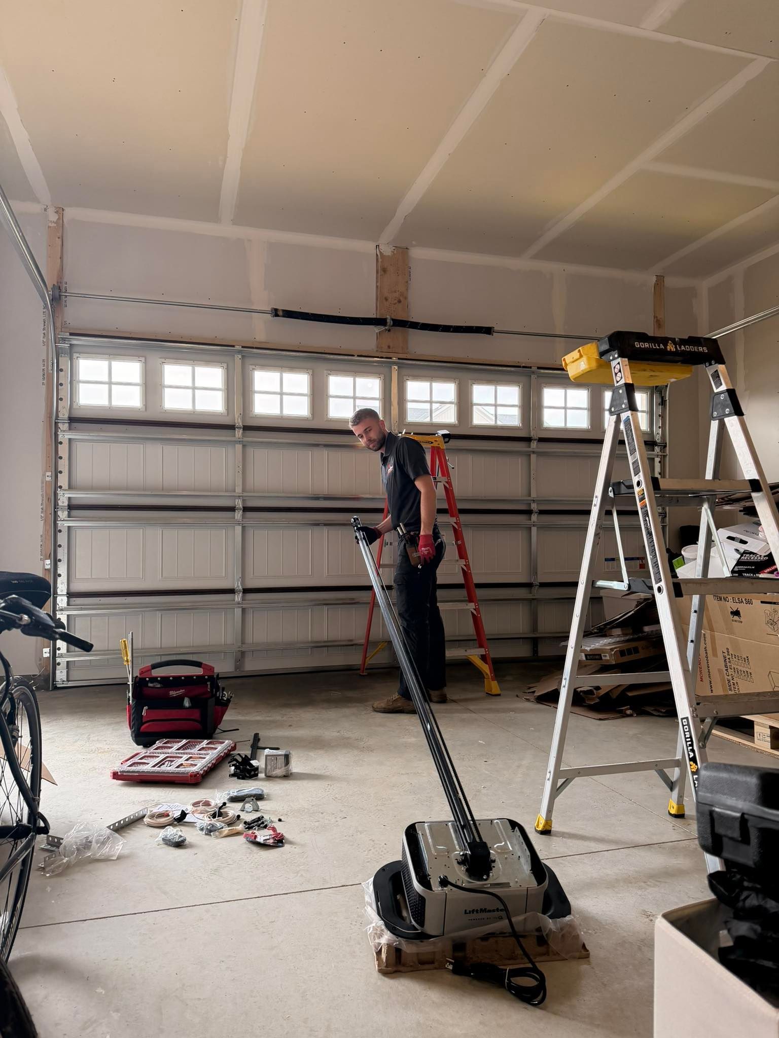 Worker in a garage using a floor grinder beside a ladder and tools.