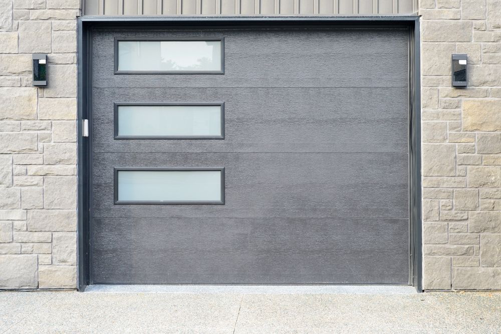 Modern gray garage door with three frosted windows, framed by stone walls.