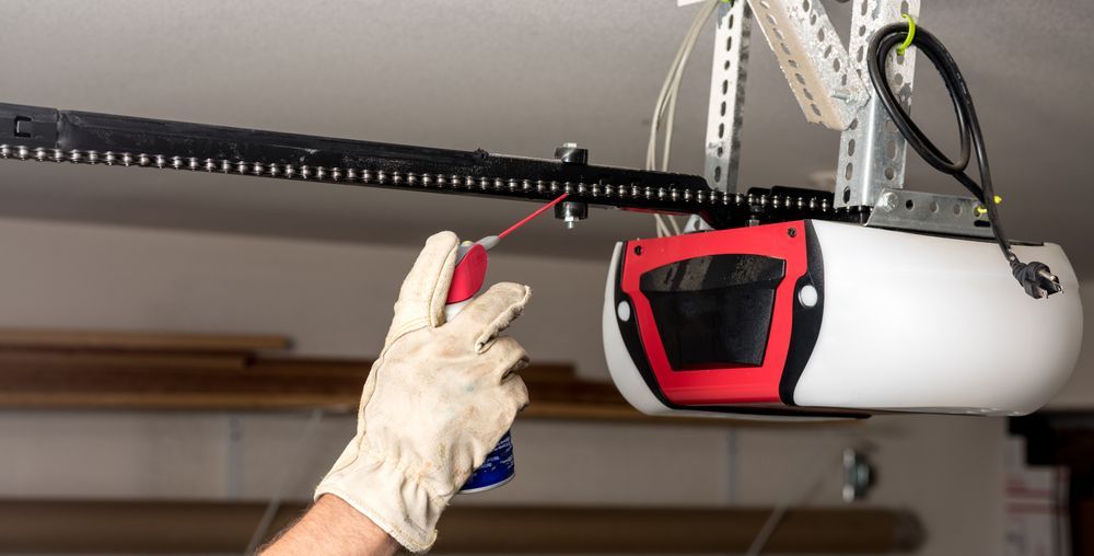 Gloved hand holding a red remote to open a garage door with a mounted opener unit