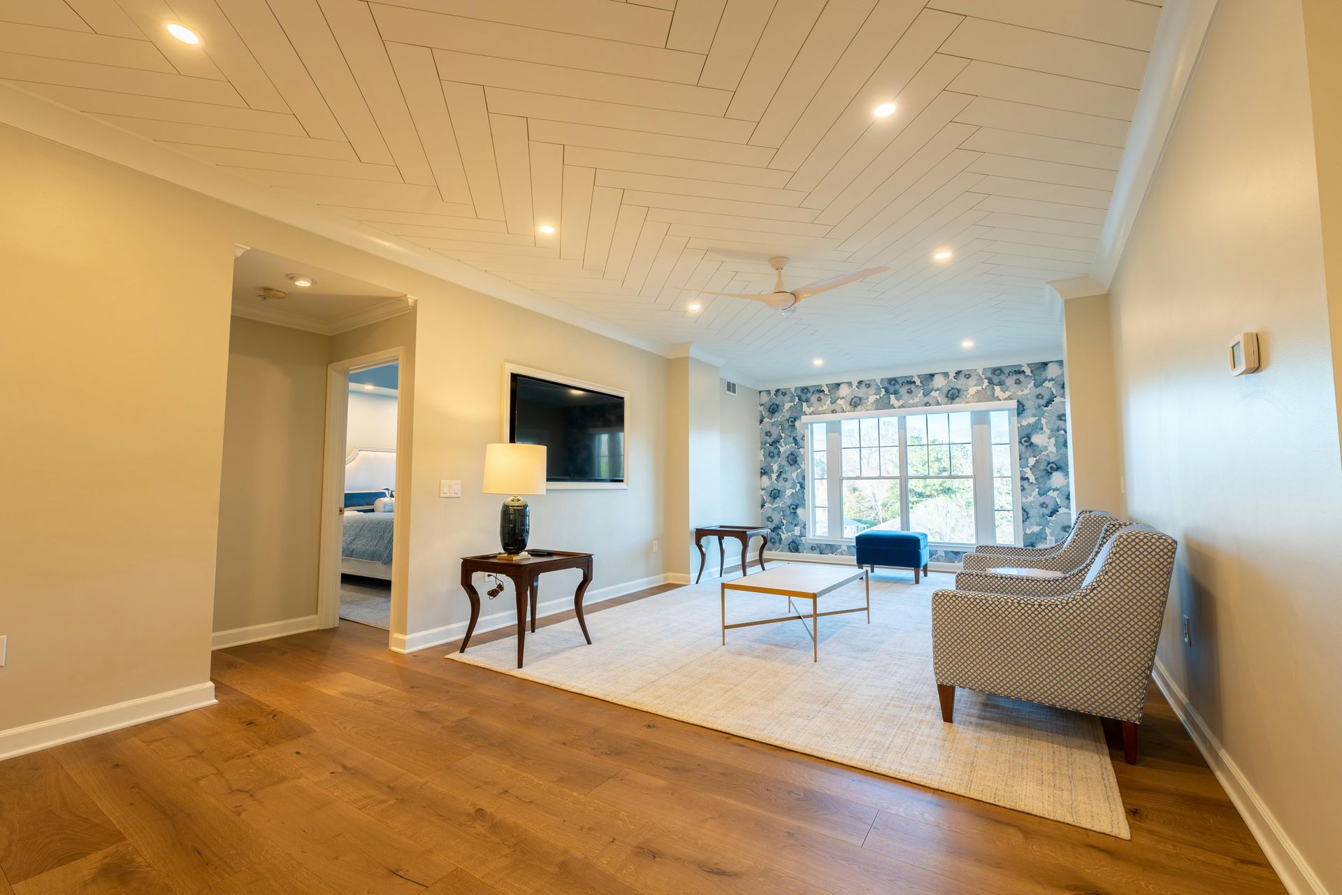 A living room with hardwood floors, a patterned ceiling, a wall-mounted TV, two chairs, and a window with blue curtains.