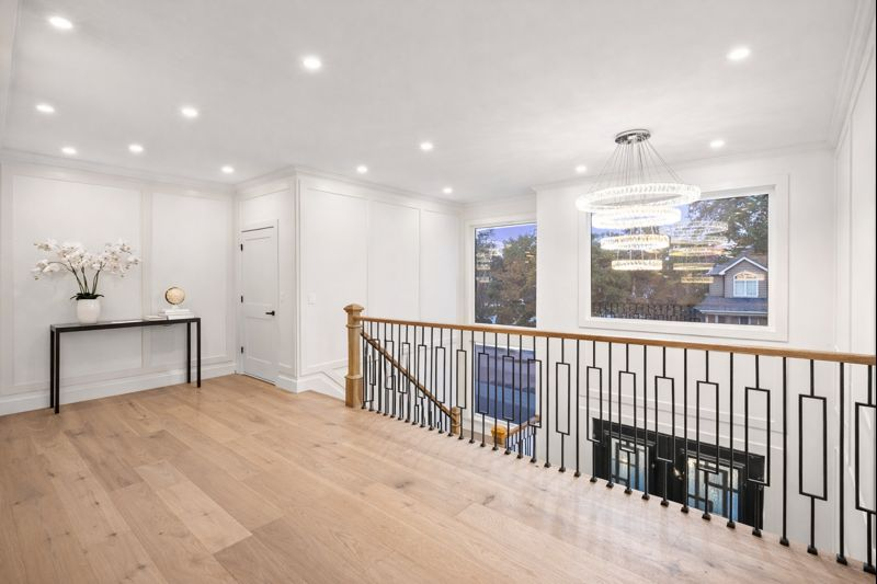 Bright upstairs hallway with wood floors, white walls, black railing, and large windows overlooking trees.