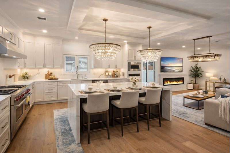 Bright open-plan kitchen and living room with white cabinets, island seating, chandelier, and fireplace