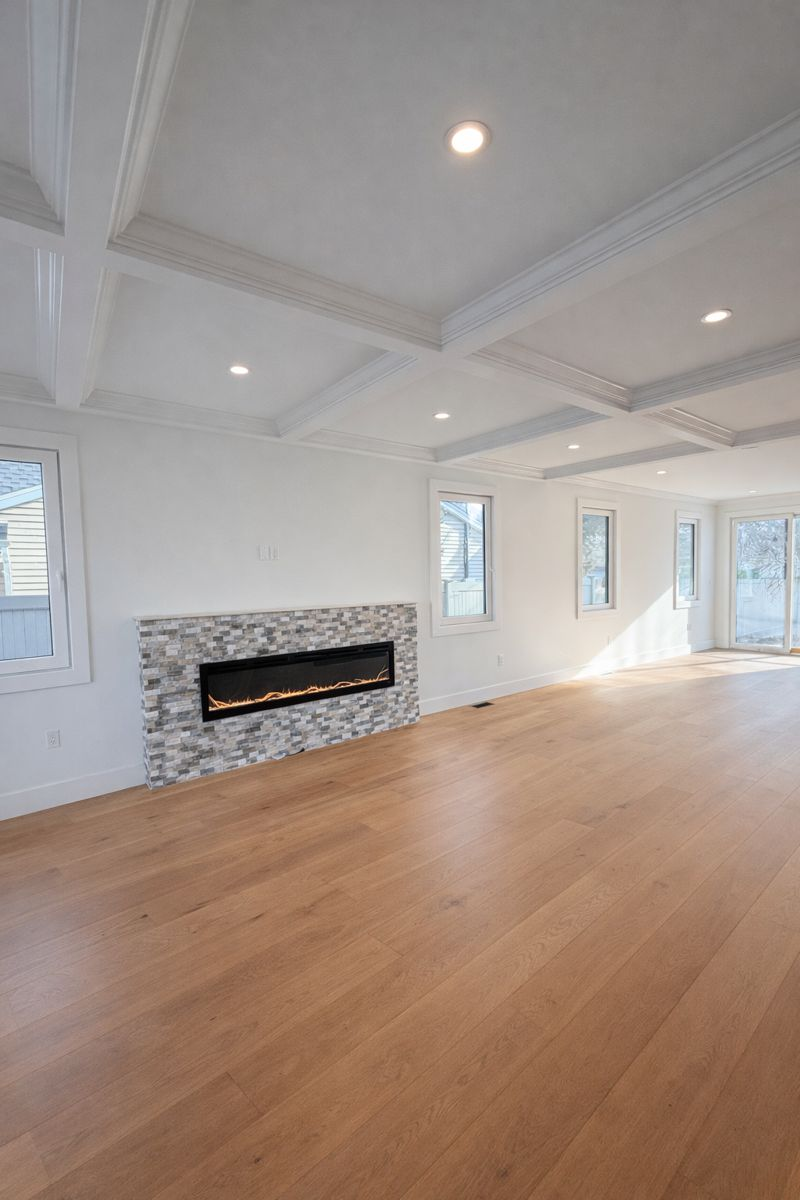Bright empty living room with wood floors, white walls, recessed lights, and a stone fireplace.