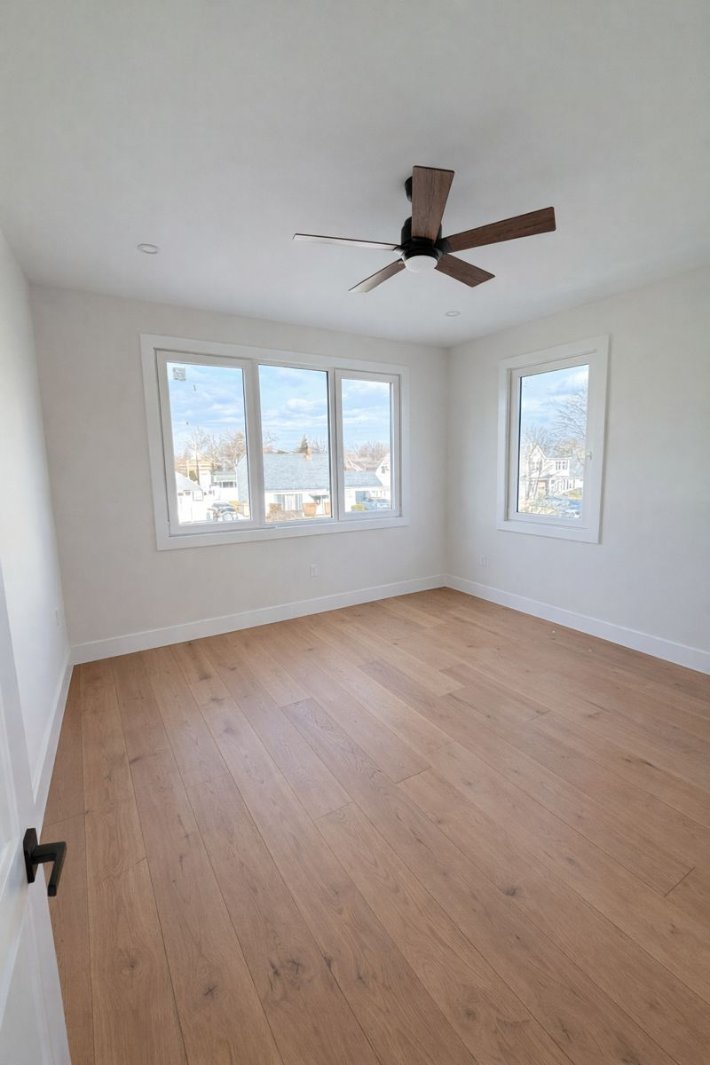 Empty bright bedroom with light wood floors, white walls, three windows, and a ceiling fan