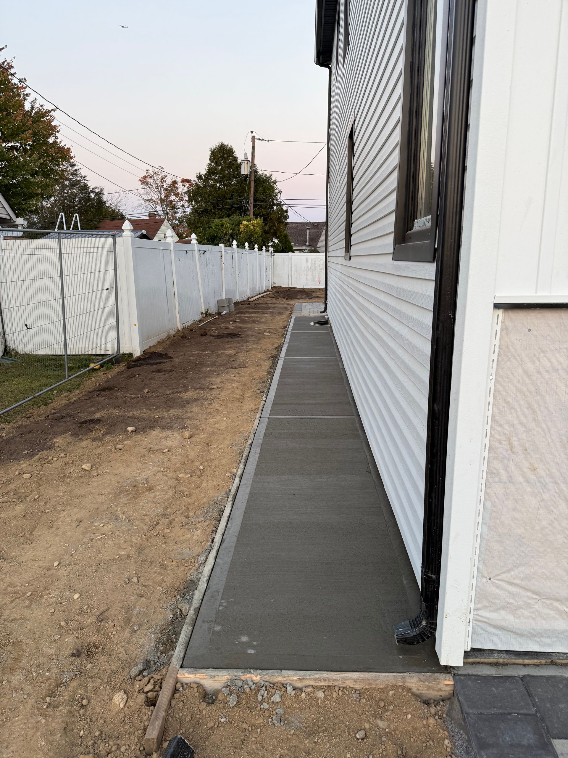 Narrow concrete walkway beside a white house and fence in a dirt side yard