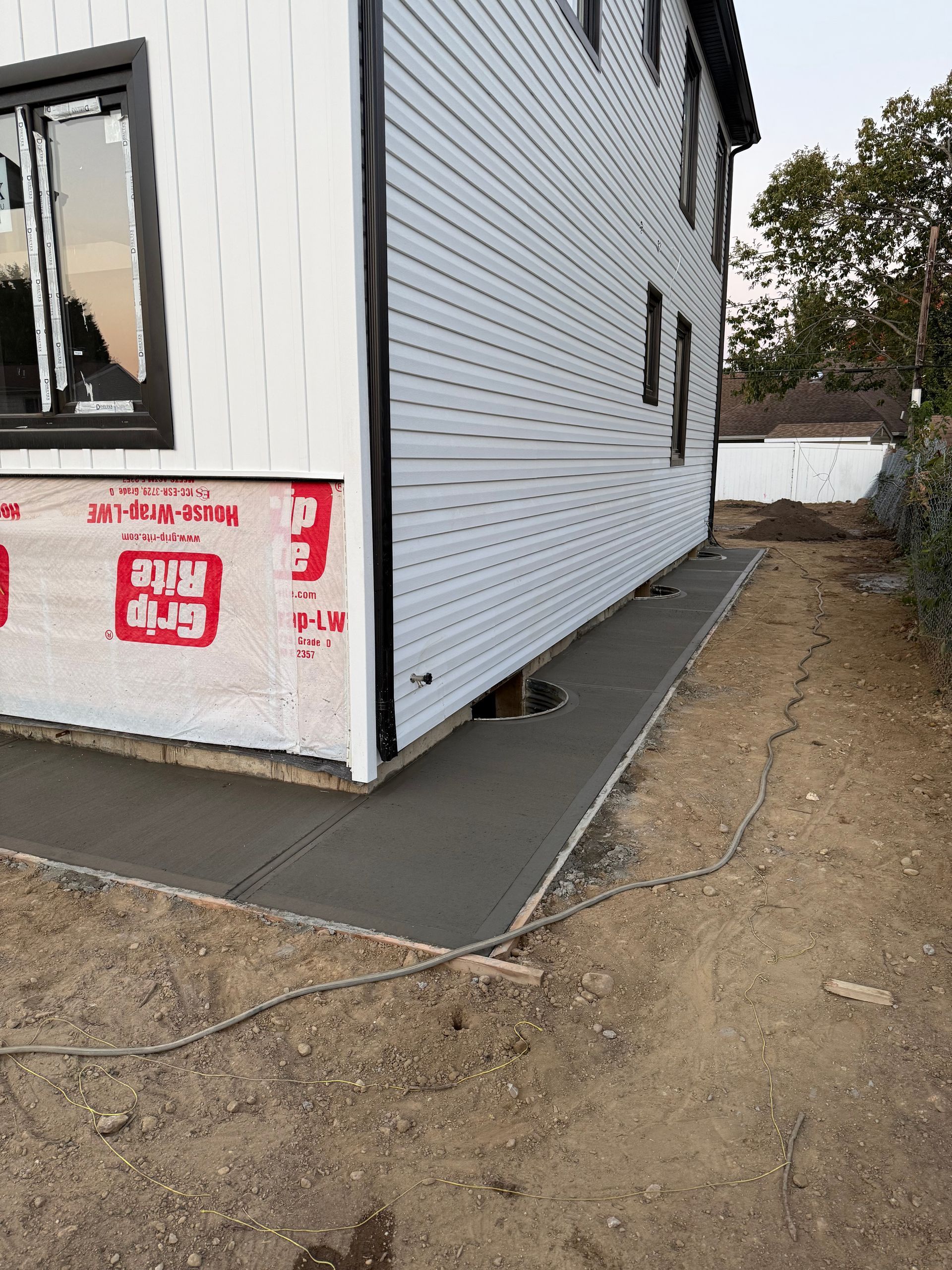 Side of a white house on a concrete slab beside a dirt yard, with a window and red construction notice.