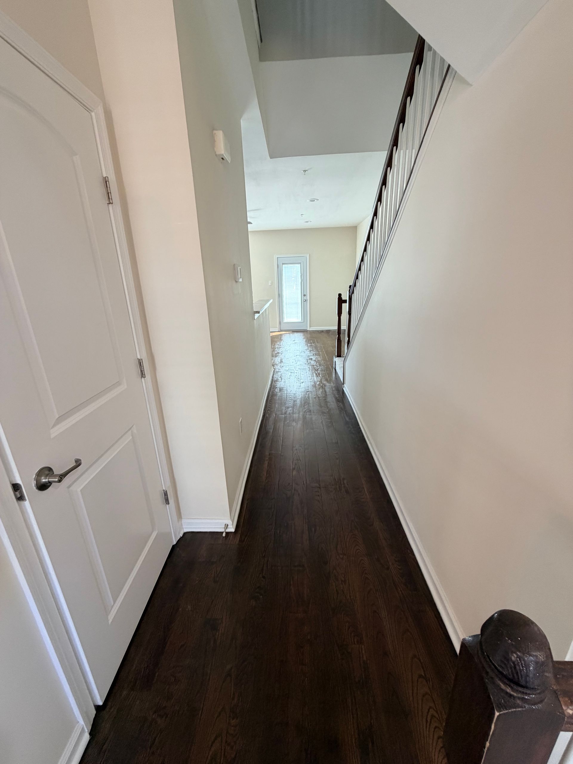 Narrow hallway with dark wood floor, white walls, staircase railing, and a bright doorway at the end