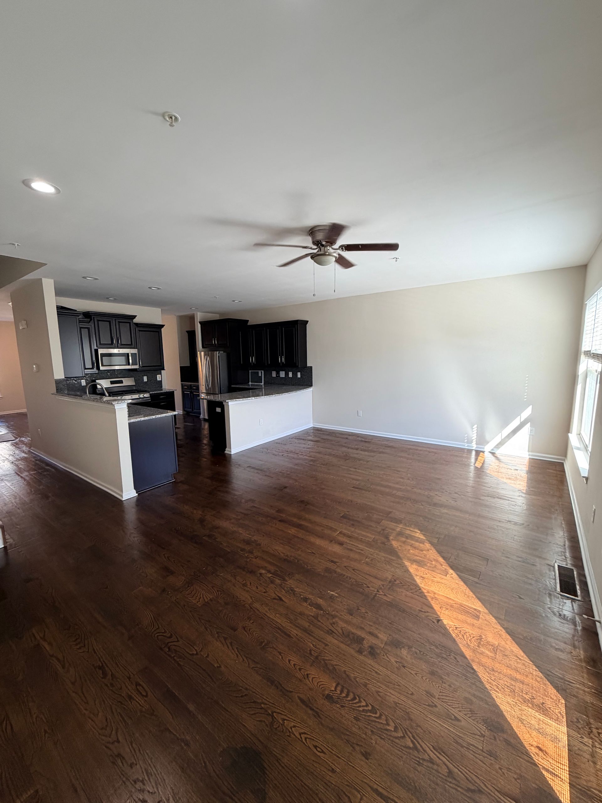 Empty living room with dark wood floors, white walls, ceiling fan, and bright sunlight from the right.