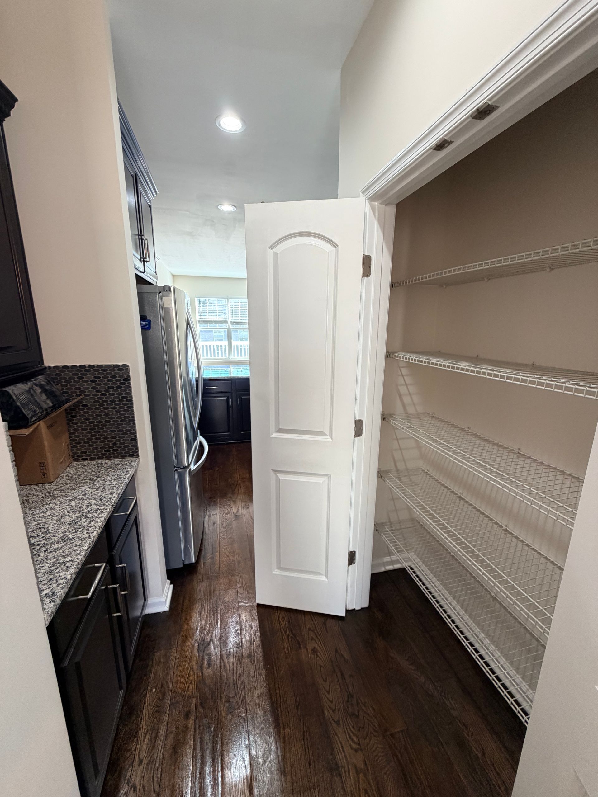 Narrow pantry hallway with empty shelves on the right, granite counter on the left, and a white door at the end.