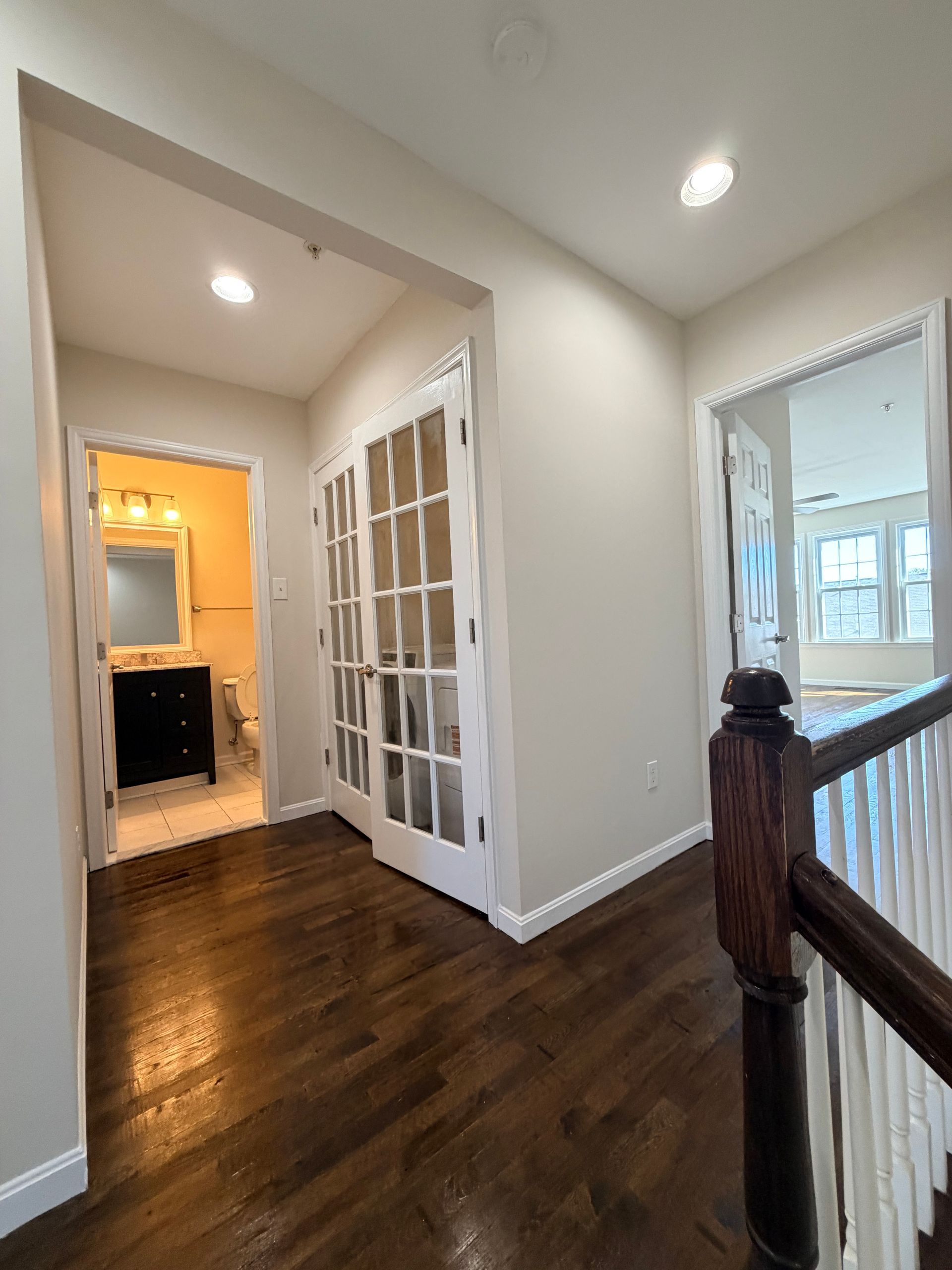 Hallway with dark hardwood floors, white walls, French doors, and a bathroom visible through an open doorway.