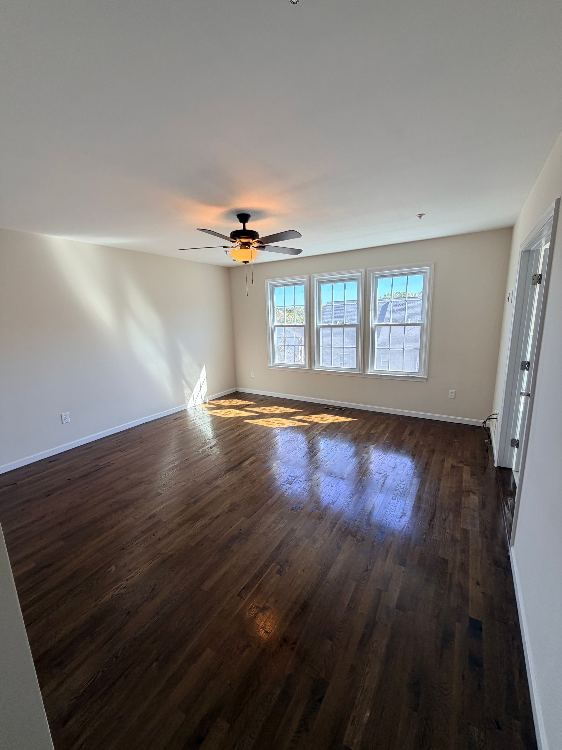 Empty living room with dark wood floors, ceiling fan, and sunlight from three windows.