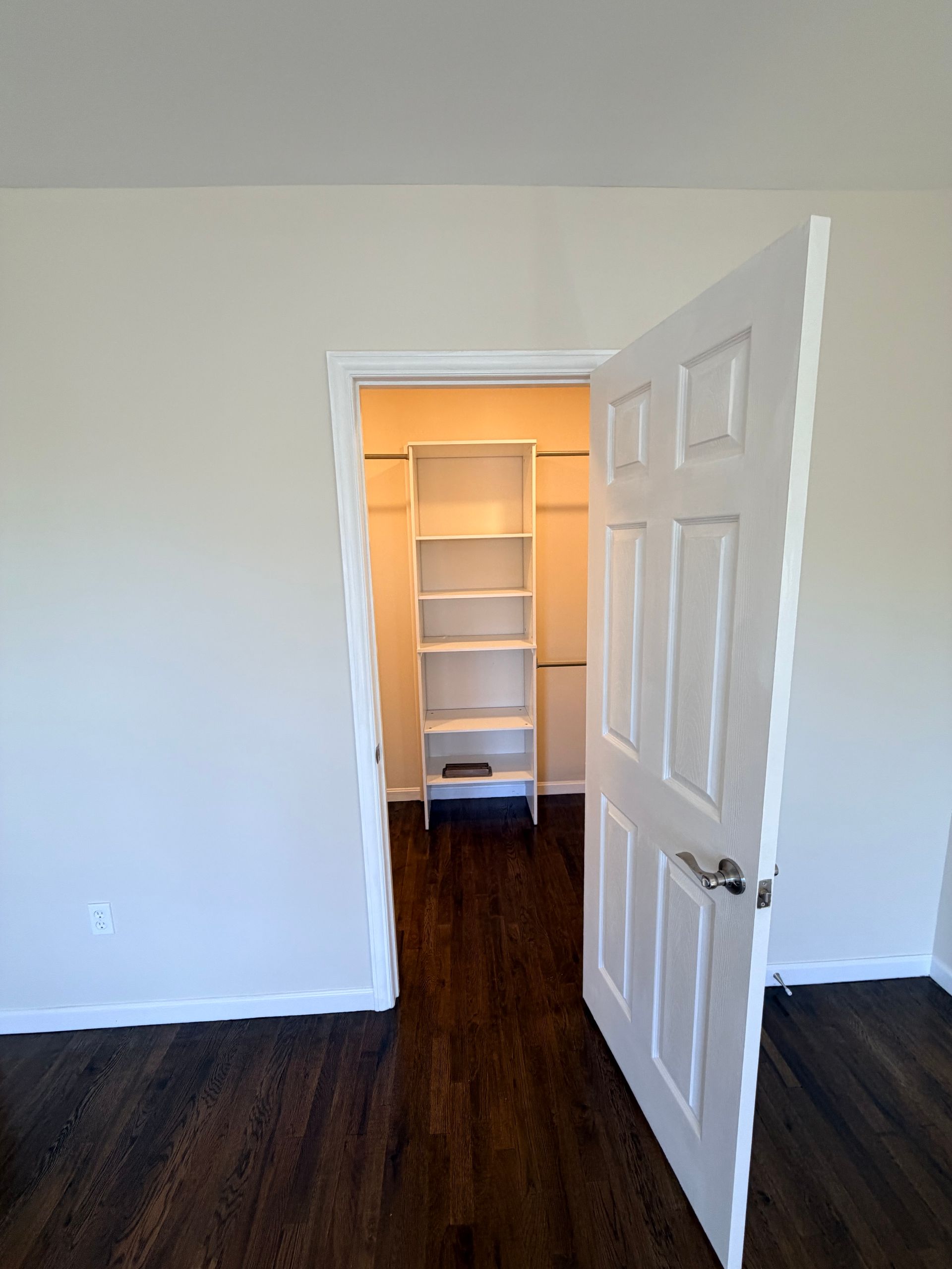 Open door to a closet with white shelves in a room with dark wood floors and white walls