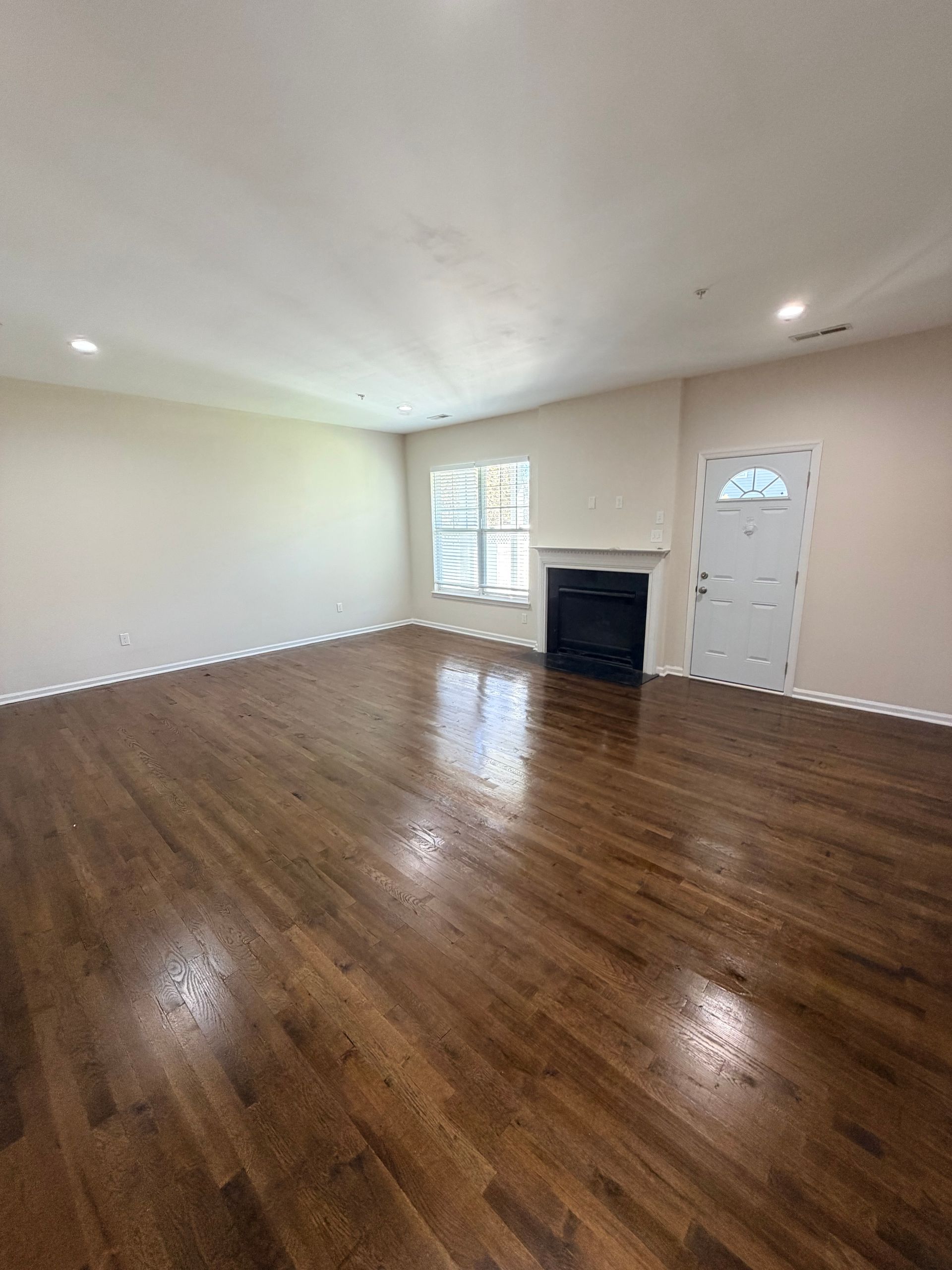 Empty living room with hardwood floors, white walls, a fireplace, and a front door by a window.