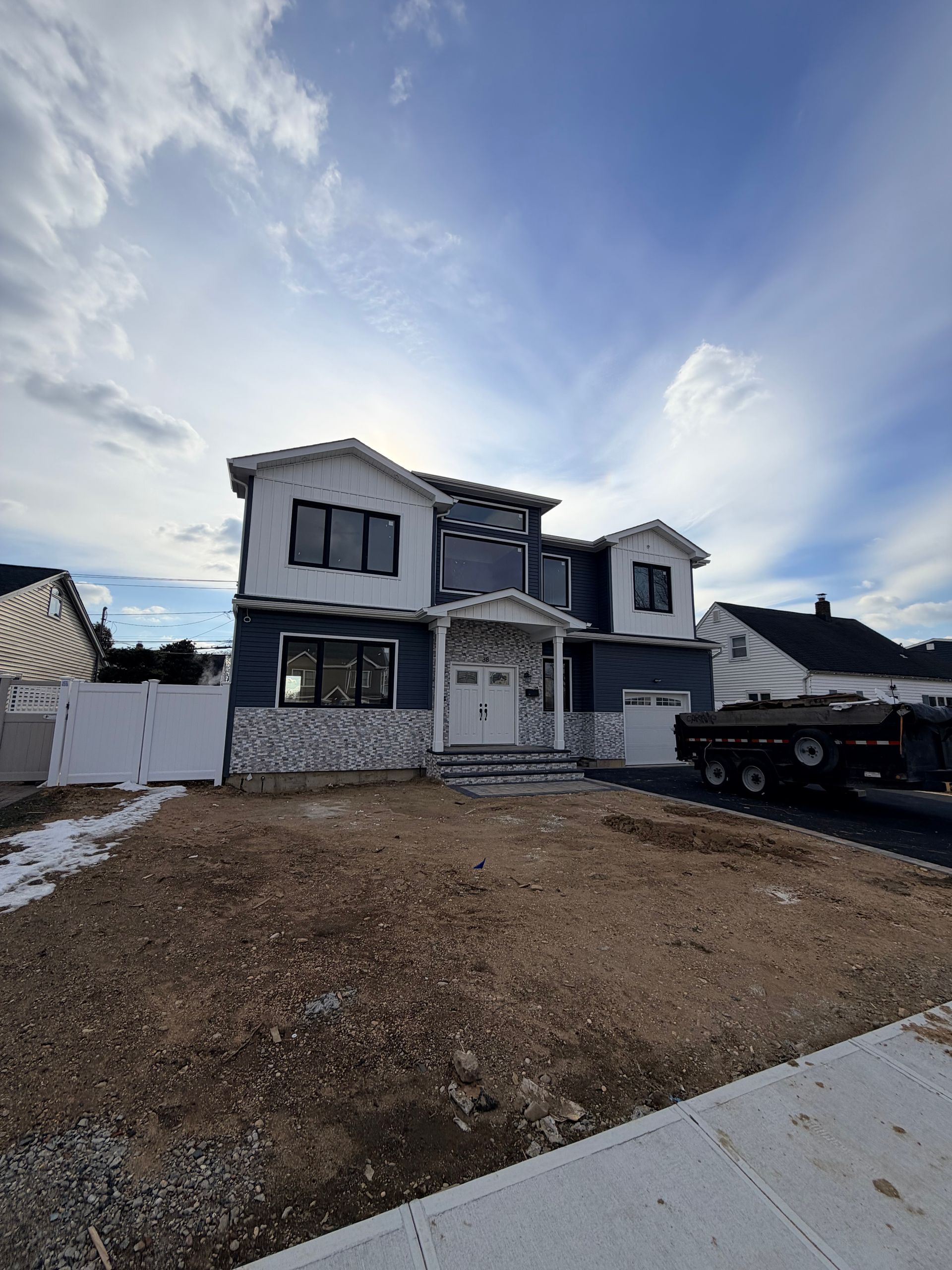 Two-story house under construction with gravel driveway and a flatbed trailer parked beside it