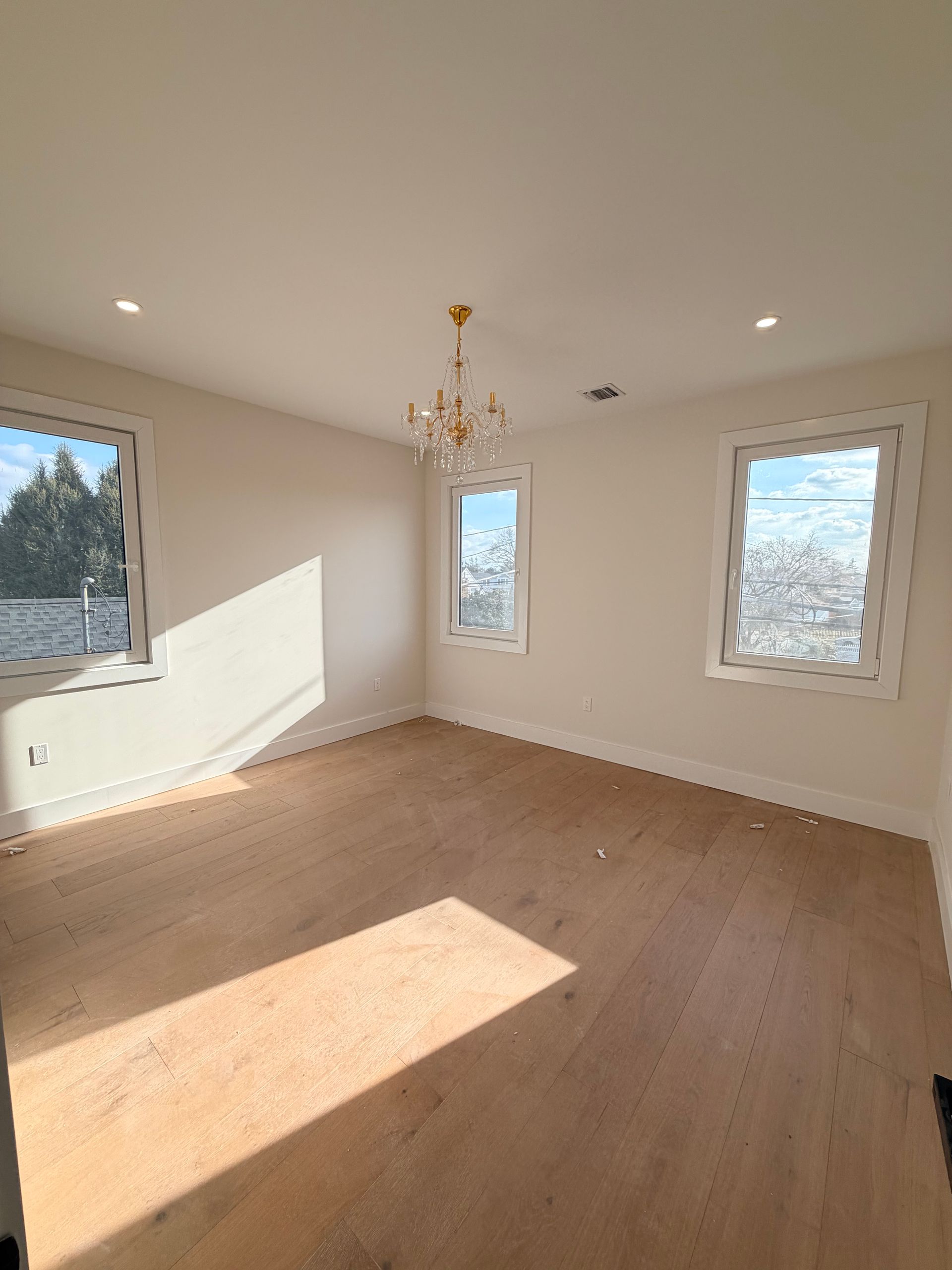 Empty sunlit room with hardwood floors, beige walls, and a small chandelier near three windows.