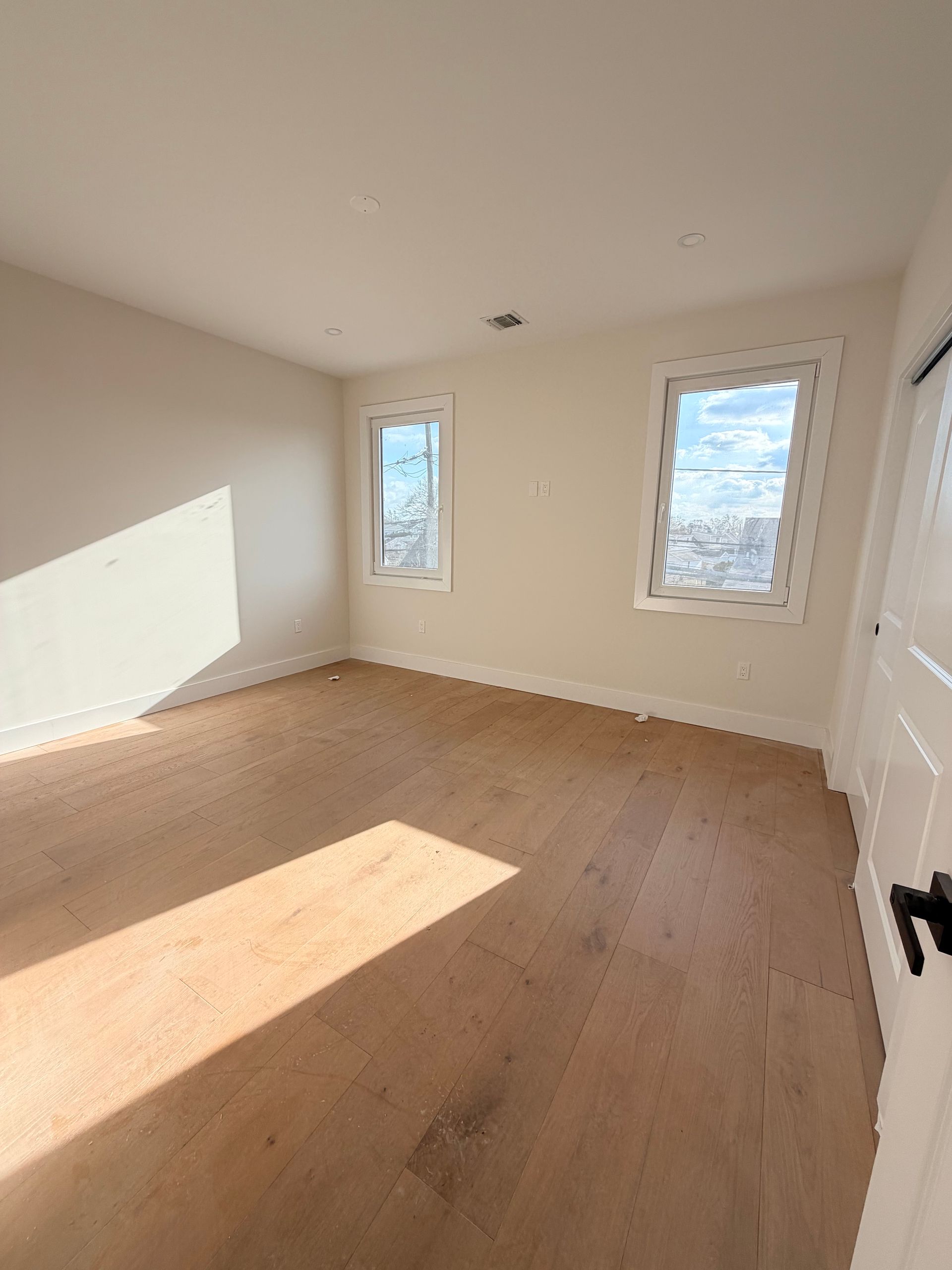 Empty sunlit room with light wood floors, white walls, and two windows.