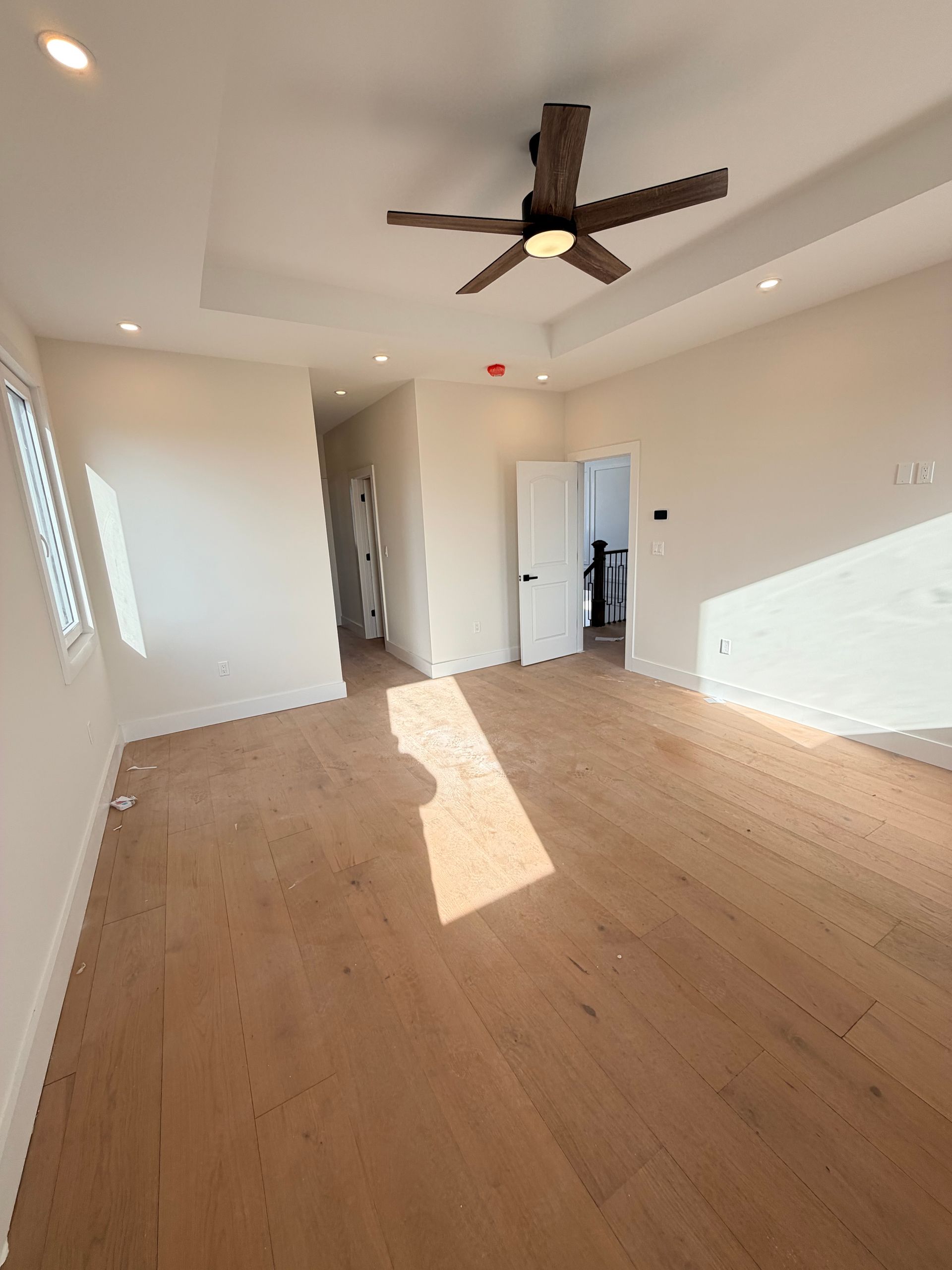 Empty modern living room with wood floors, ceiling fan, white walls, and sunlight streaming in through a window.