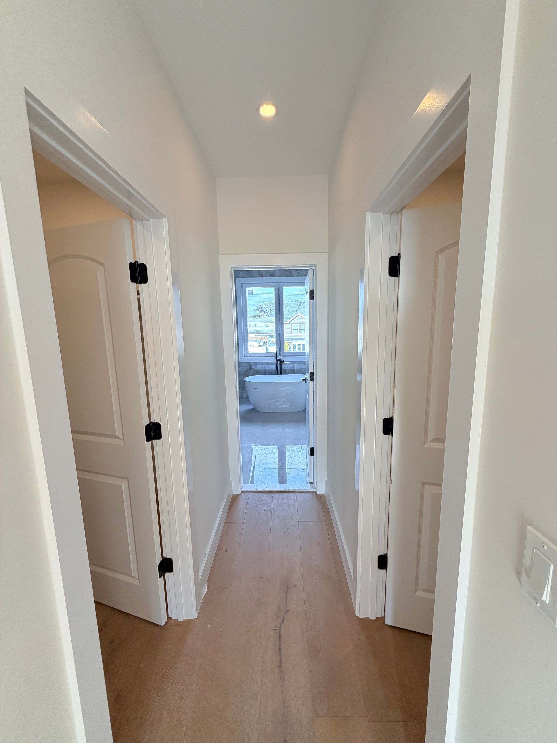 Narrow white hallway with open doors on both sides, wood floor, and a bright room visible at the end.