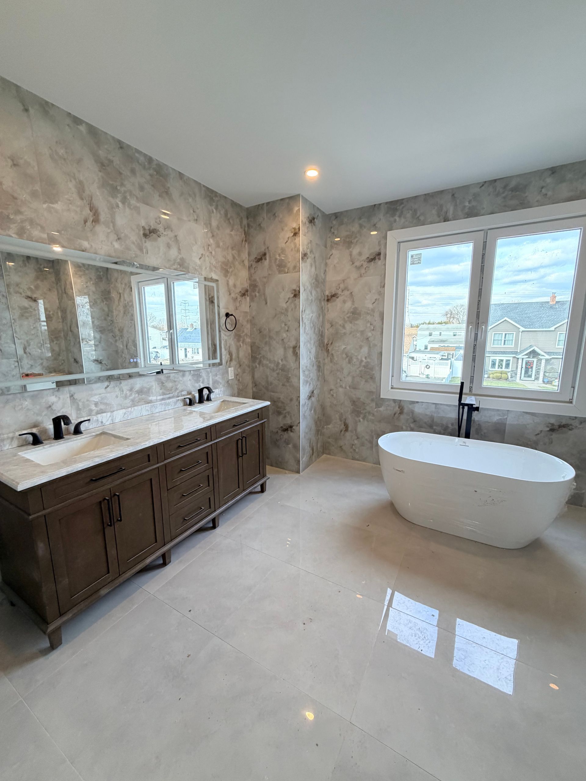 Modern bathroom with marble walls, double vanity, large mirror, freestanding tub, and window view
