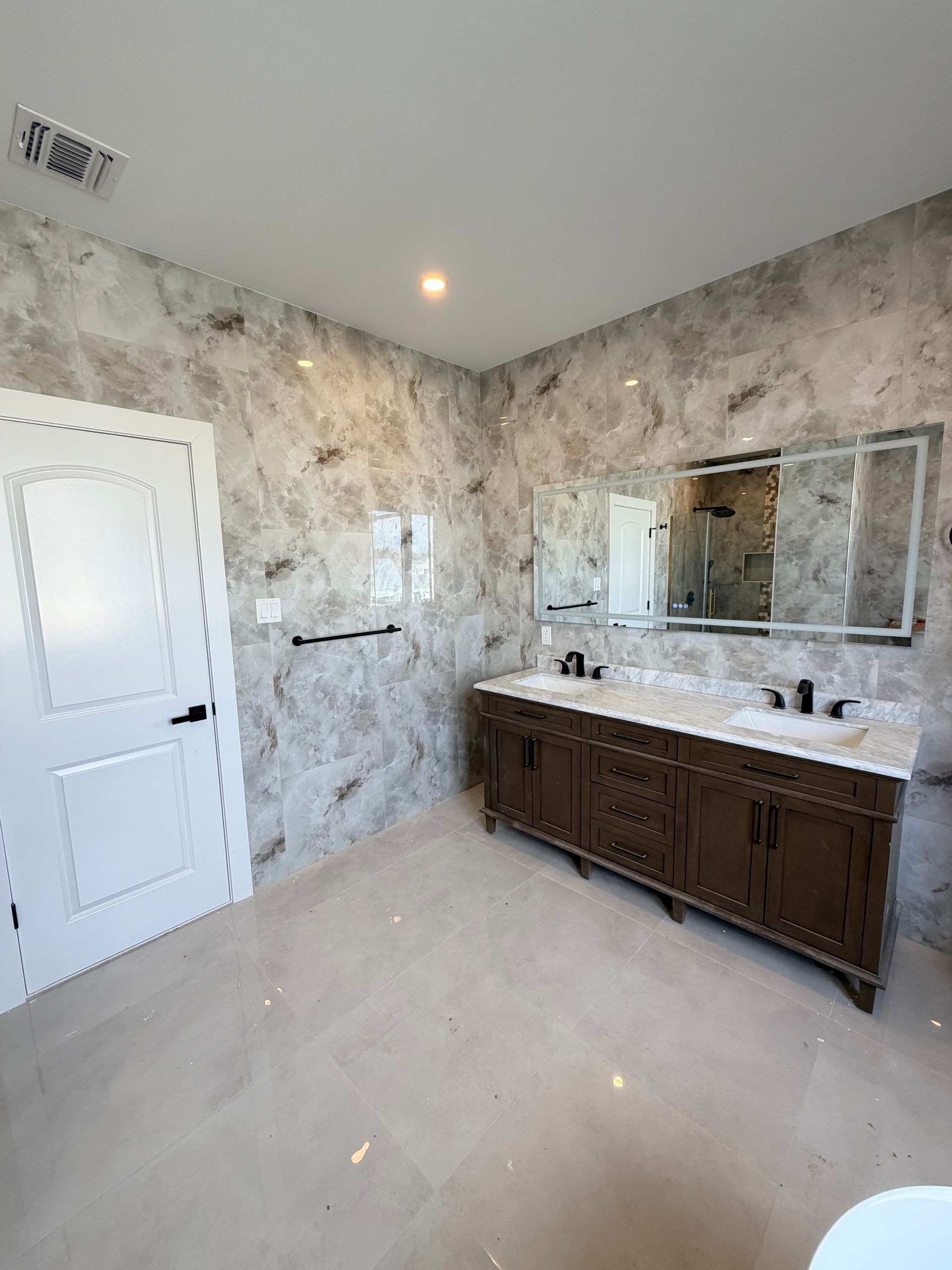 Empty marble bathroom with double vanity, large mirror, white door, and light tile floor.