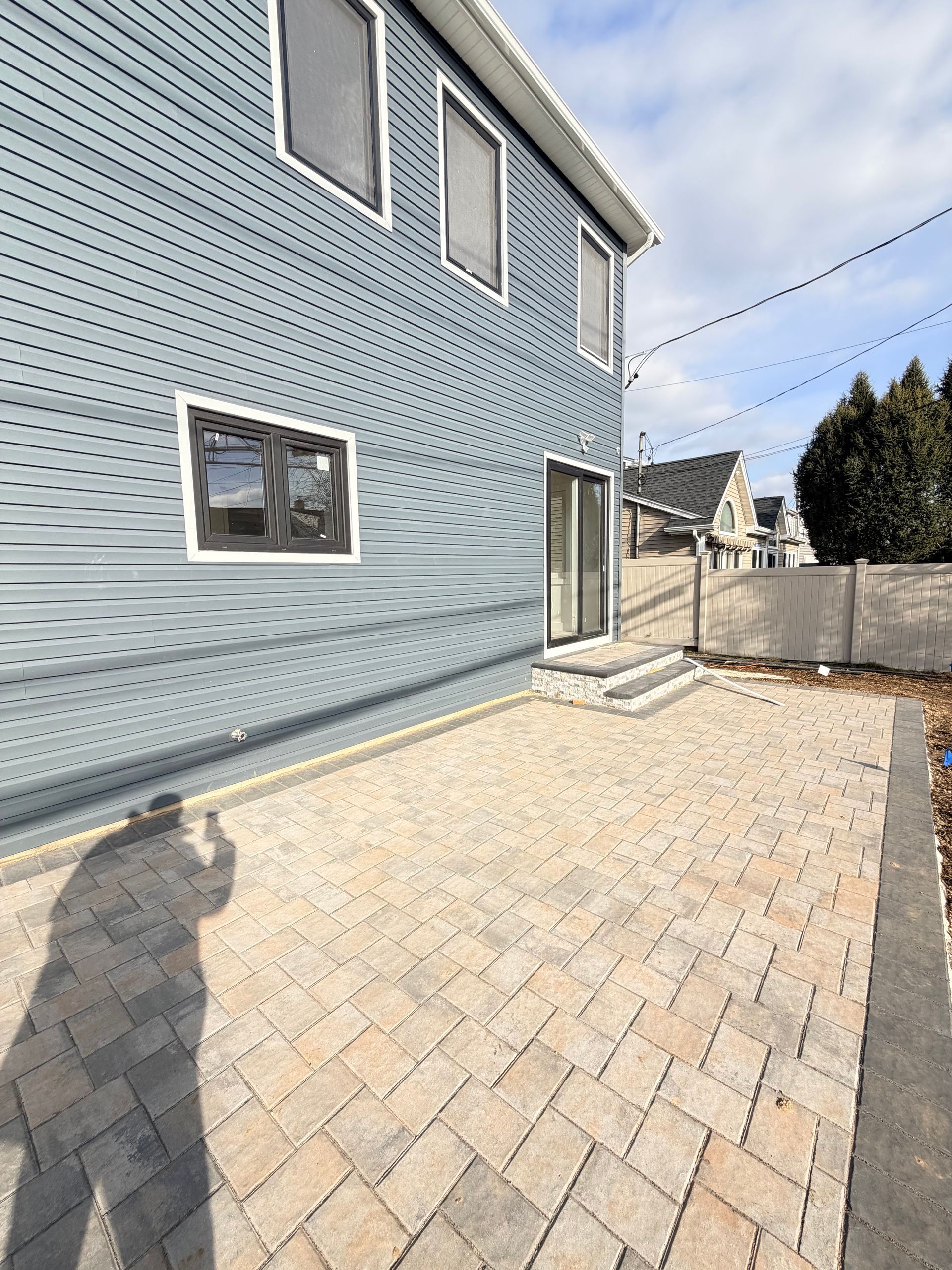 Pavered backyard beside blue house with small patio, gate, and privacy fence under bright sunlight