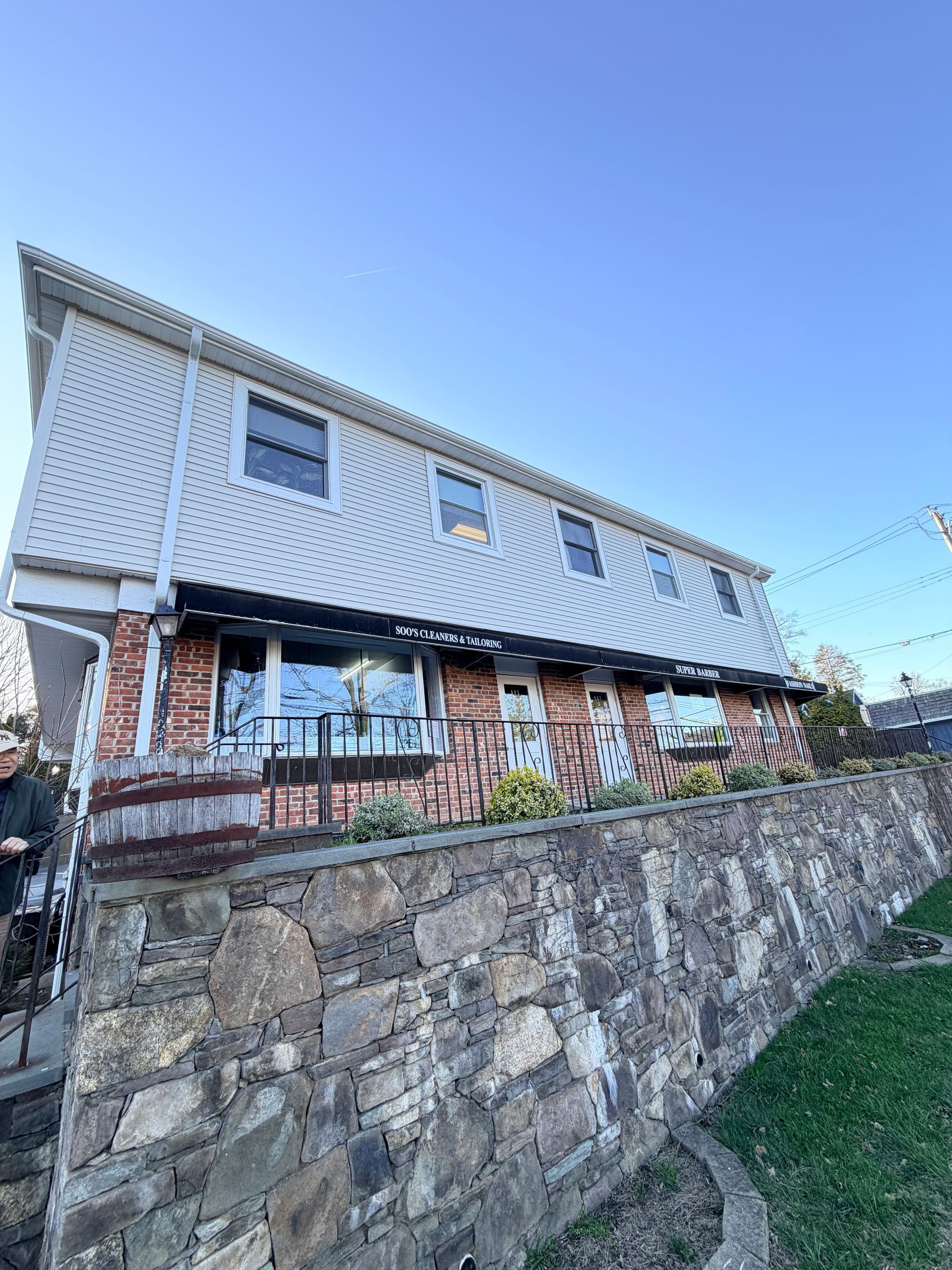 Stone house with white upper siding, brick lower level, and a sloped stone retaining wall under a blue sky