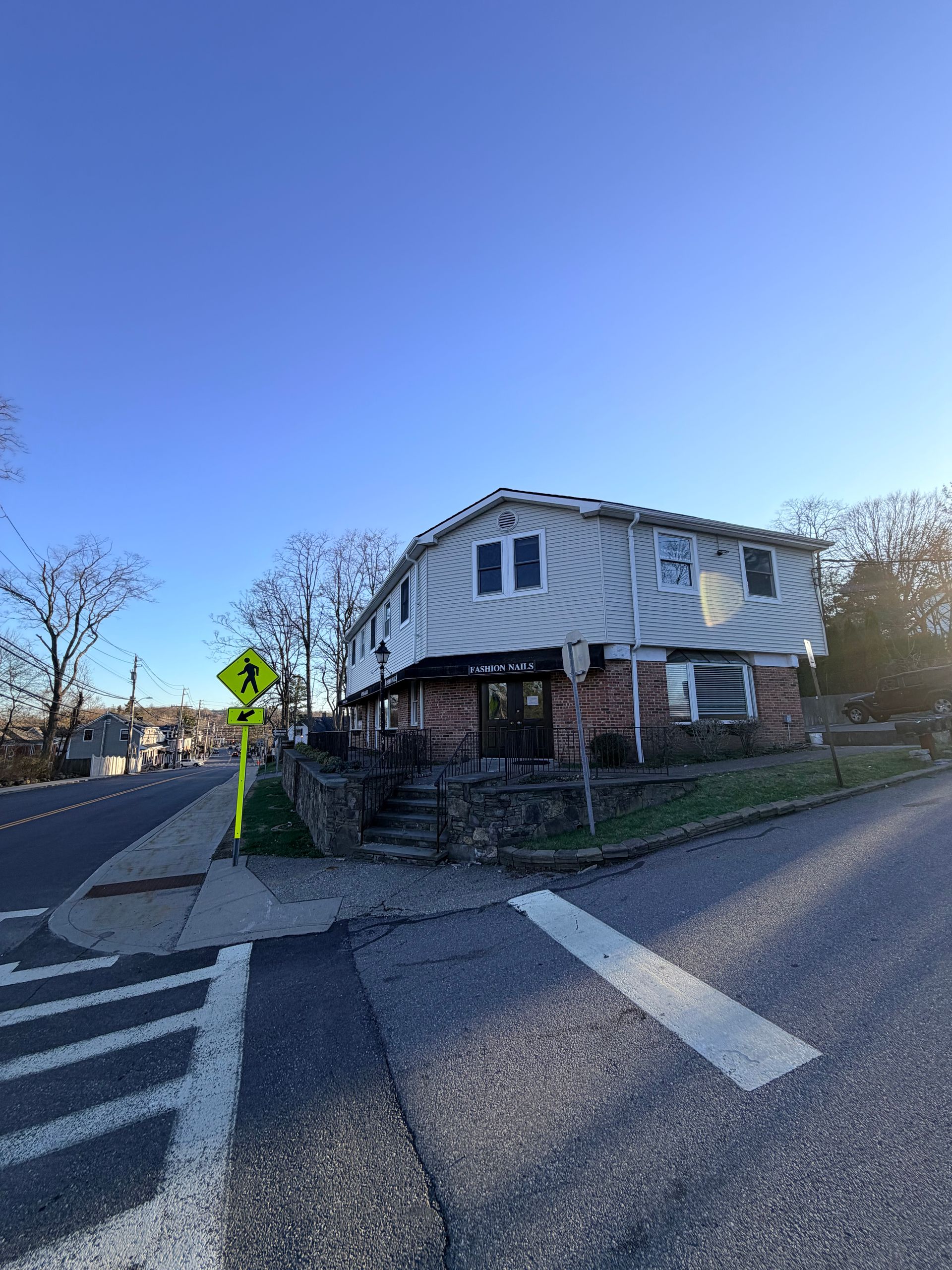 Street corner with a white house, crosswalk markings, and a pedestrian sign under a clear blue sky
