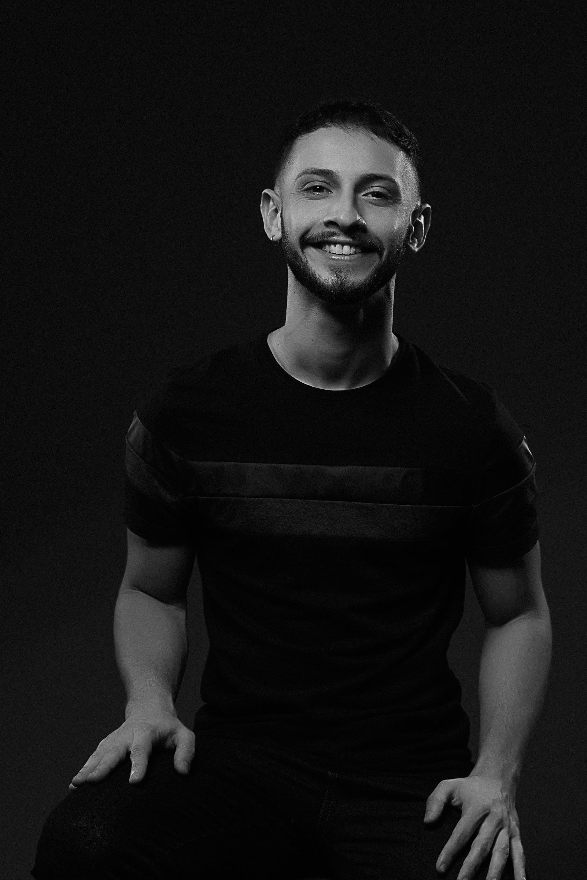 Uma pessoa sorridente, com barba curta e vestindo uma camiseta escura, está ajoelhada contra um fundo preto.
