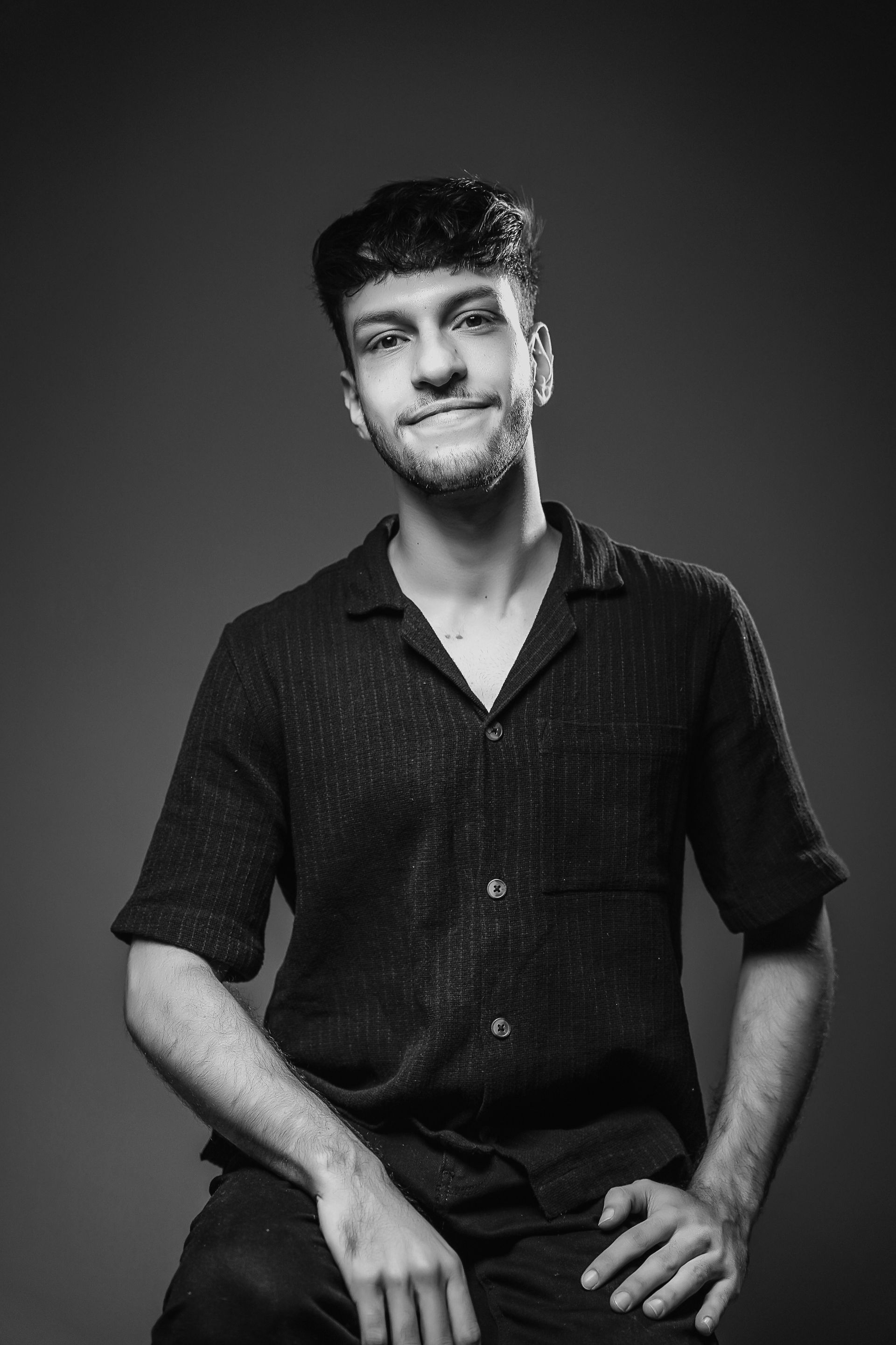 Uma pessoa com cabelo curto e escuro e barba por fazer usa uma camisa escura de botões e sorri levemente.