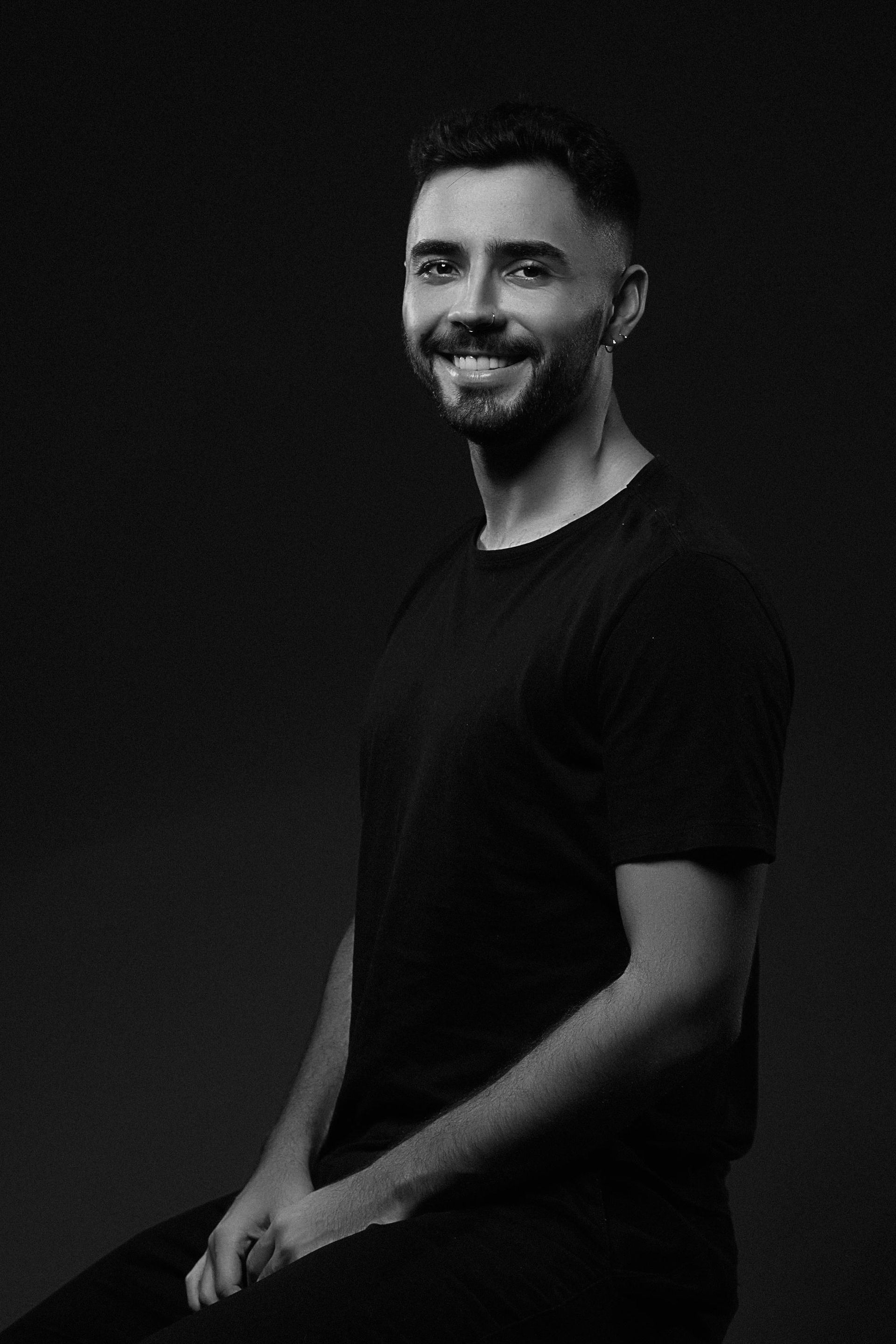 Retrato de uma pessoa sorridente com barba, vestindo uma camiseta preta contra um fundo escuro.