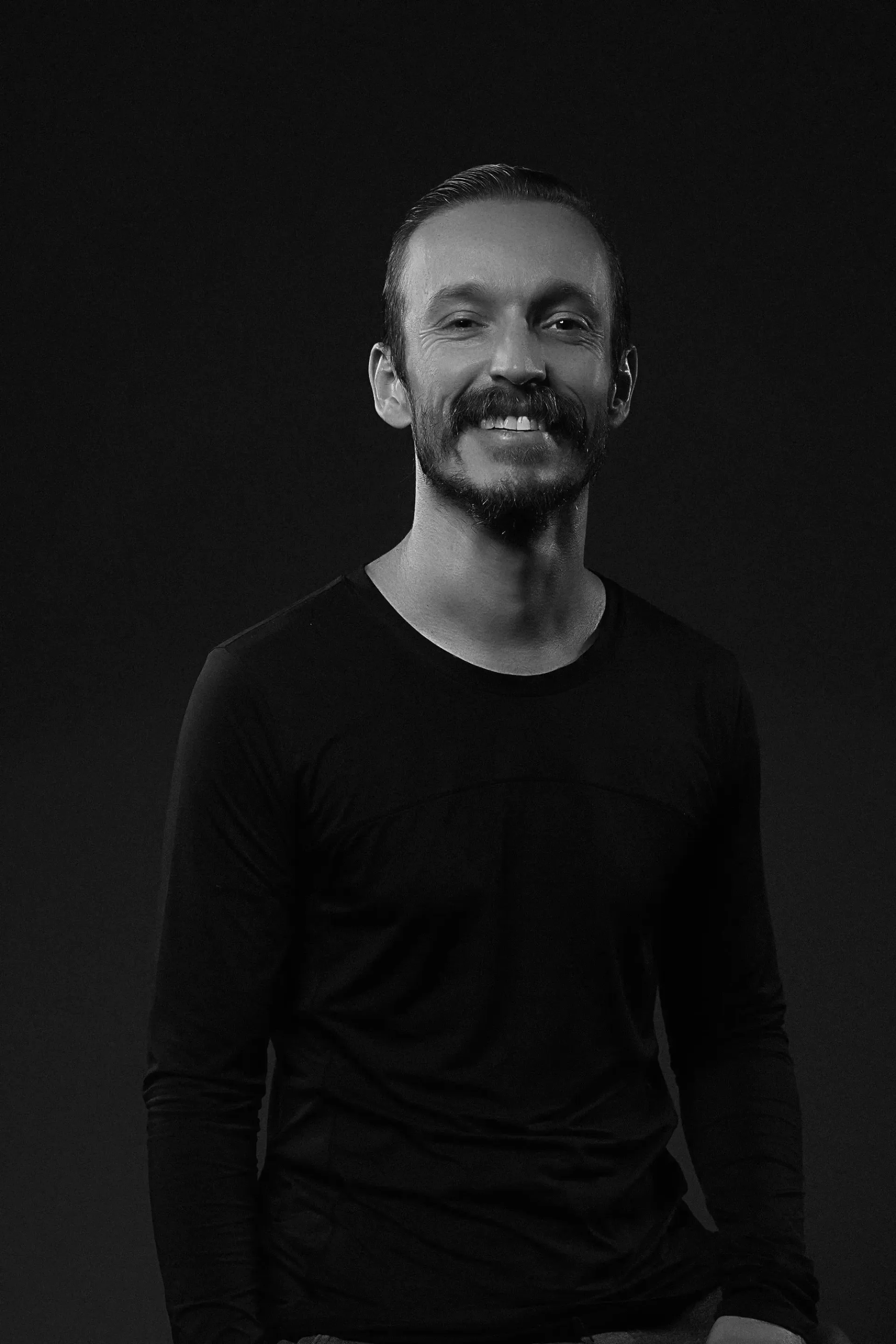 Uma pessoa sorridente, com cabelo encaracolado e barba, vestindo uma camiseta preta contra um fundo escuro.