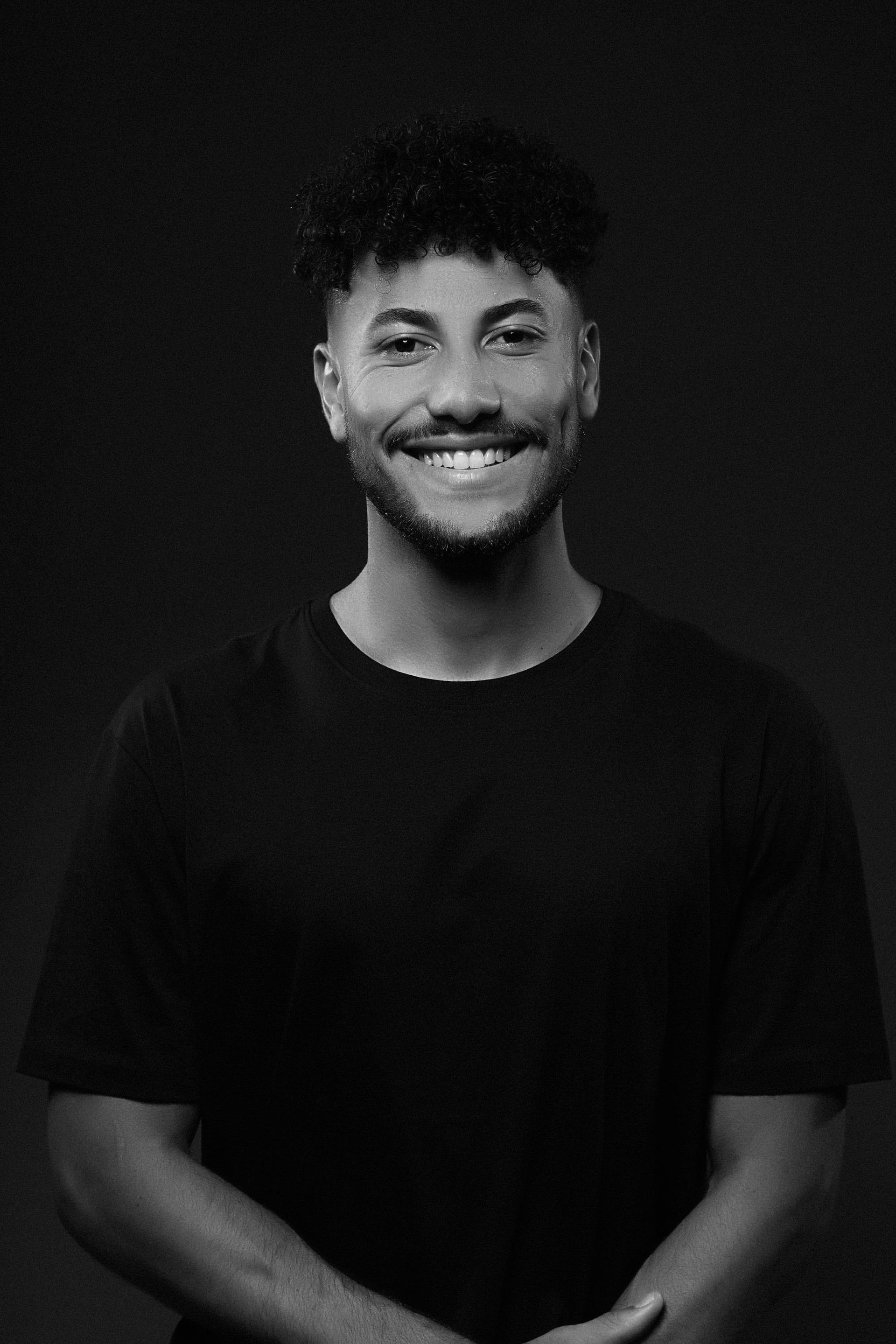 Uma pessoa sorridente, com cabelo encaracolado e barba, vestindo uma camiseta preta contra um fundo escuro.