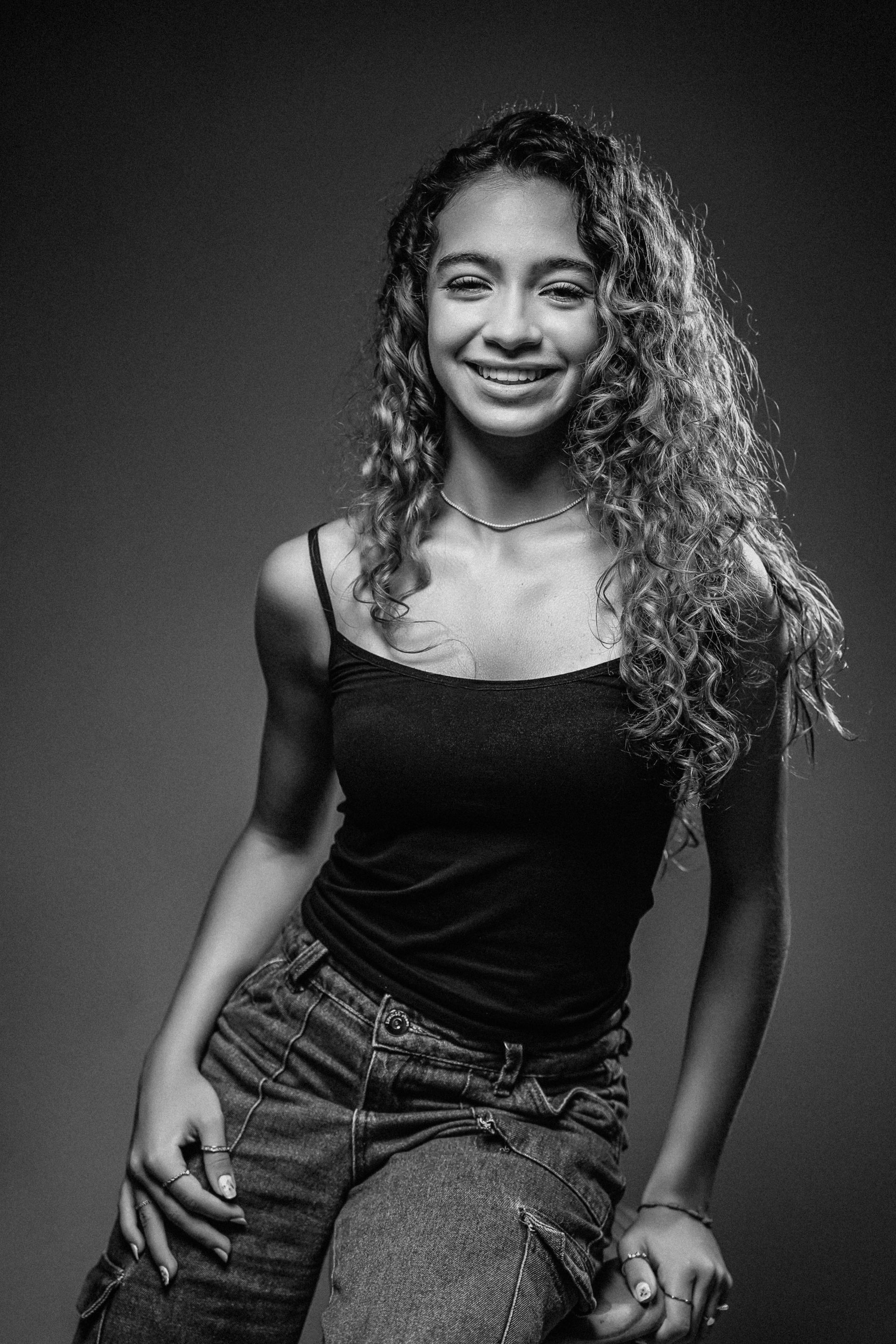 Uma pessoa com cabelos longos e cacheados sorri enquanto usa uma blusa sem mangas e calça jeans.
