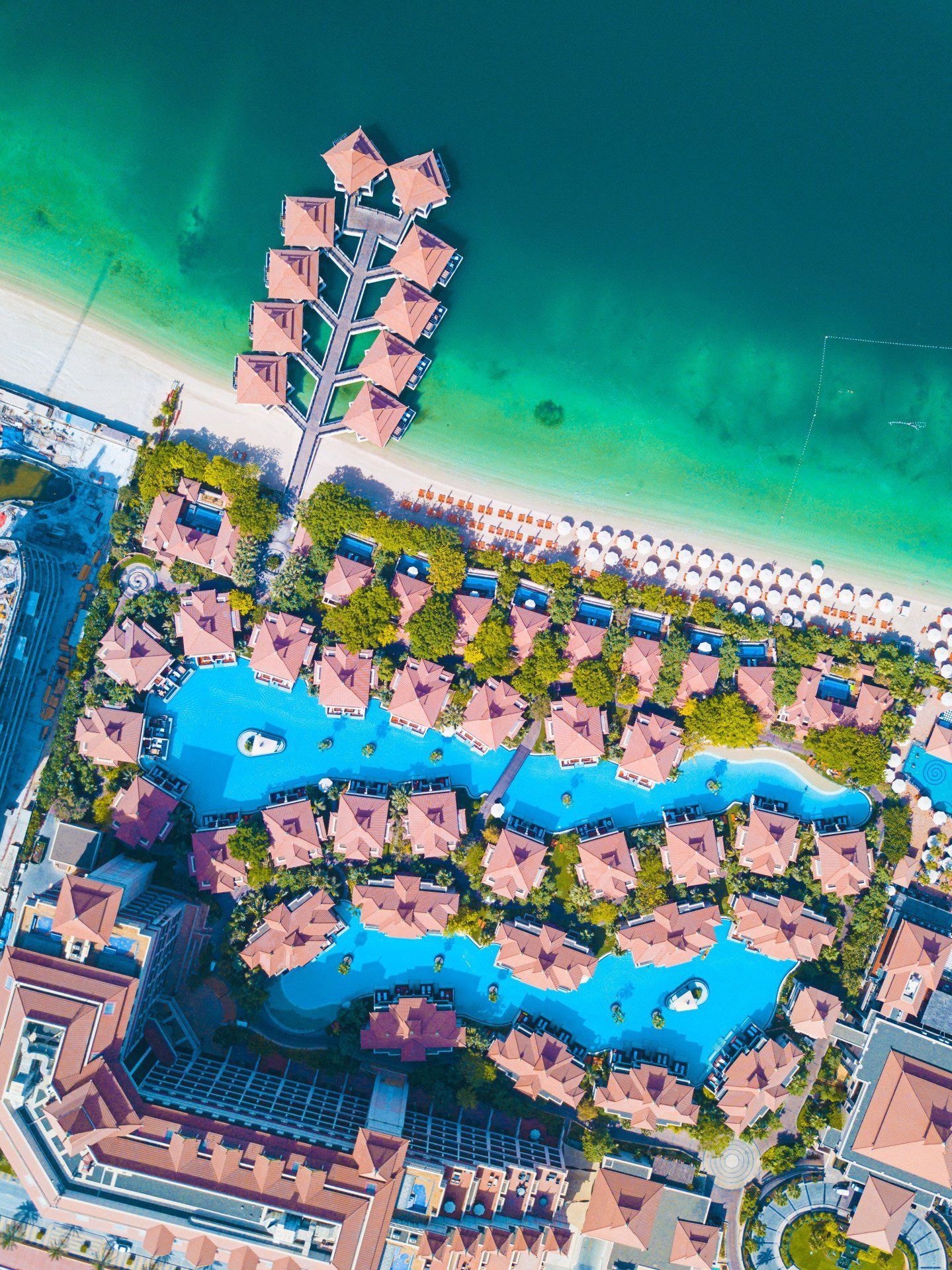 Vista aérea de um resort tropical com piscinas azuis, palmeiras e uma praia de areia ao lado de águas turquesa.