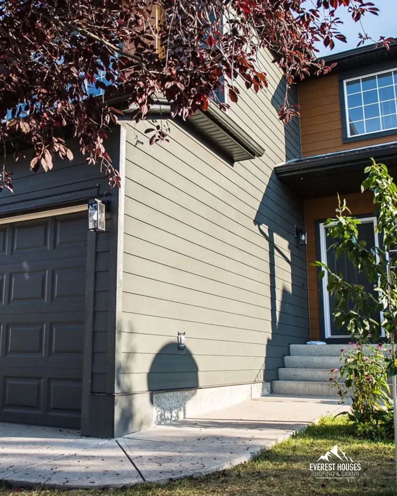 Modern house exterior with gray garage door, beige siding, and front steps under tree branches