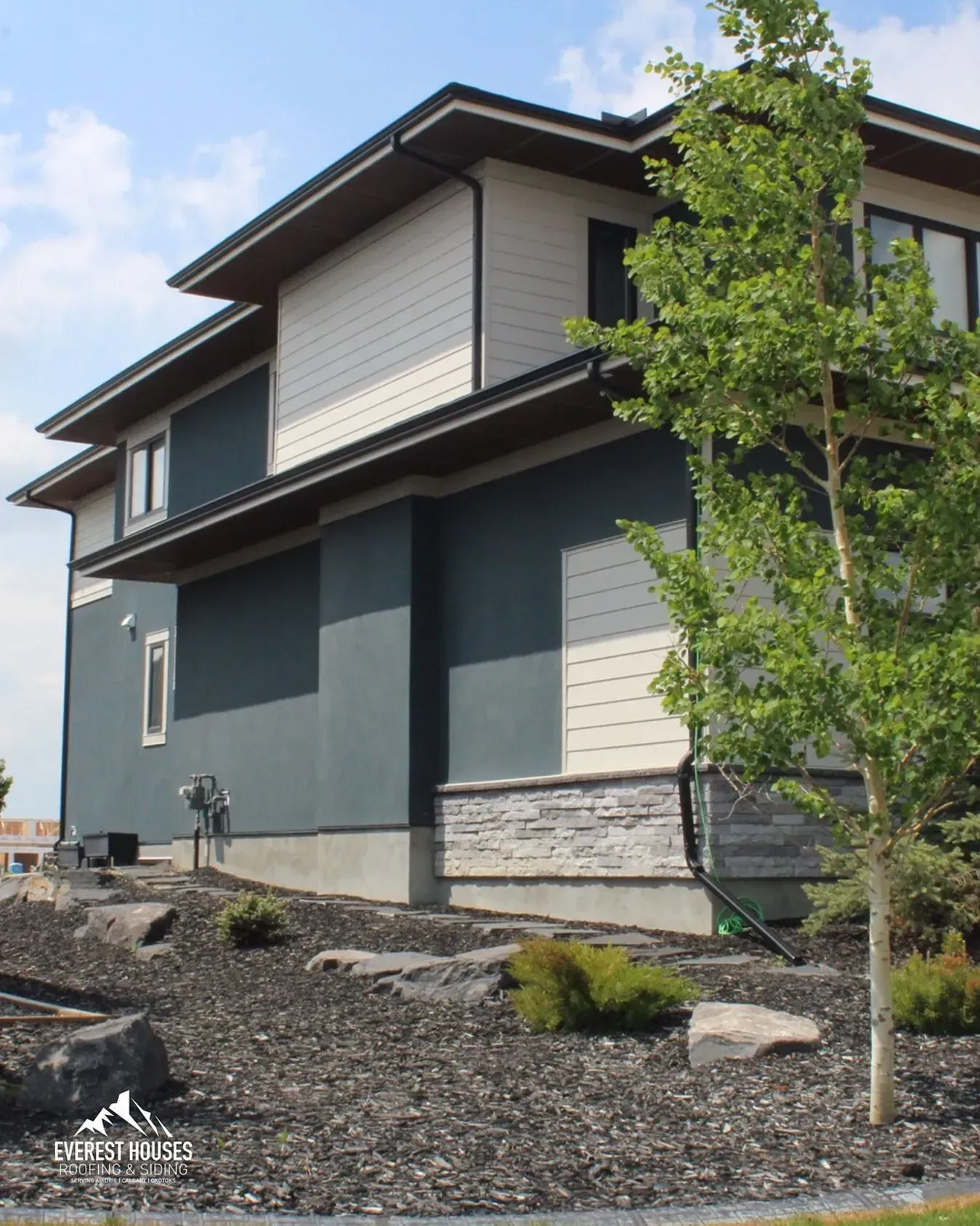Modern two-story house exterior with dark blue and white siding, gravel yard, and a young tree in front.