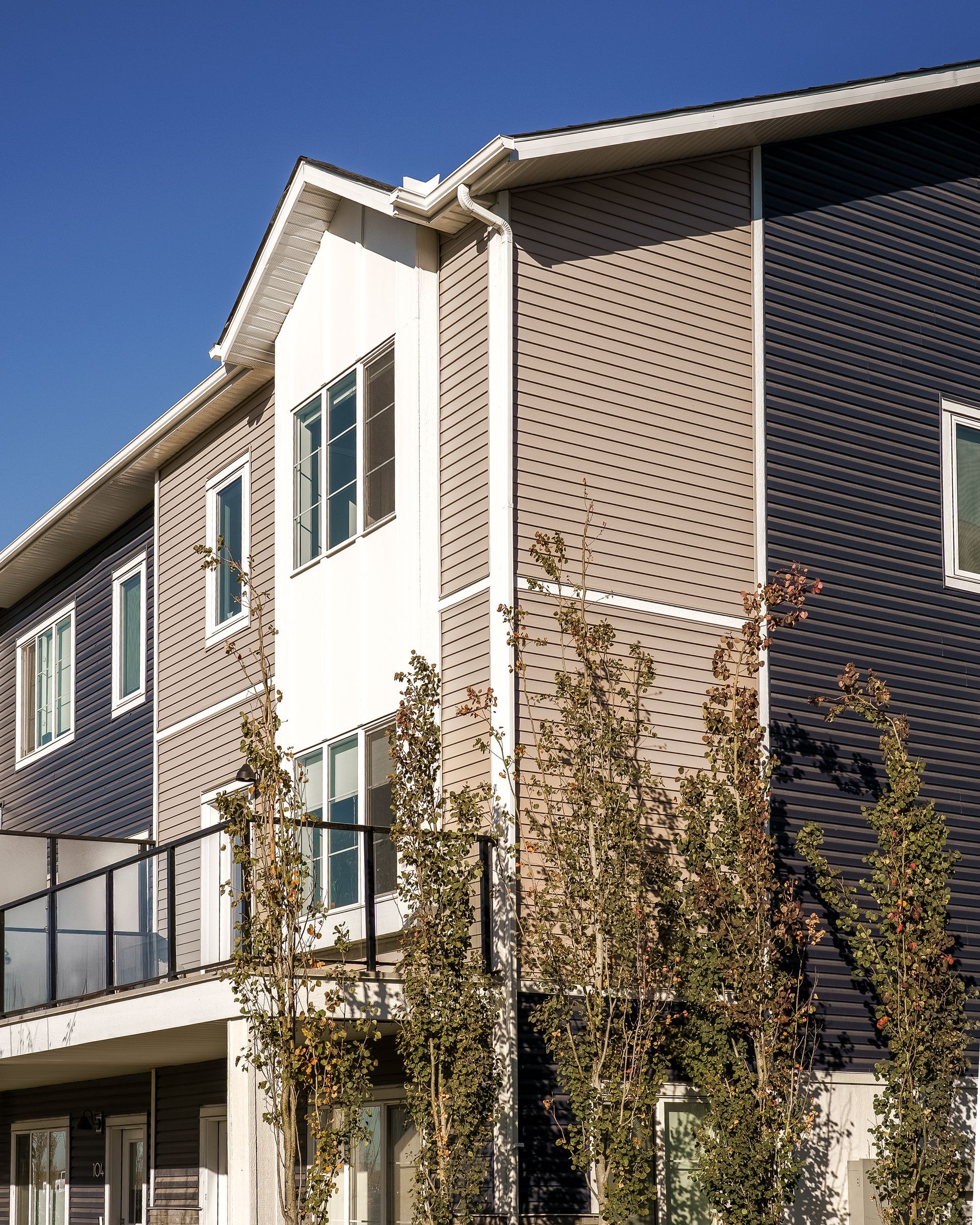 Modern apartment building with beige and dark siding, balconies, and young trees under a blue sky Modern apartment building with beige and dark siding, balconies, and young trees under a blue sky
