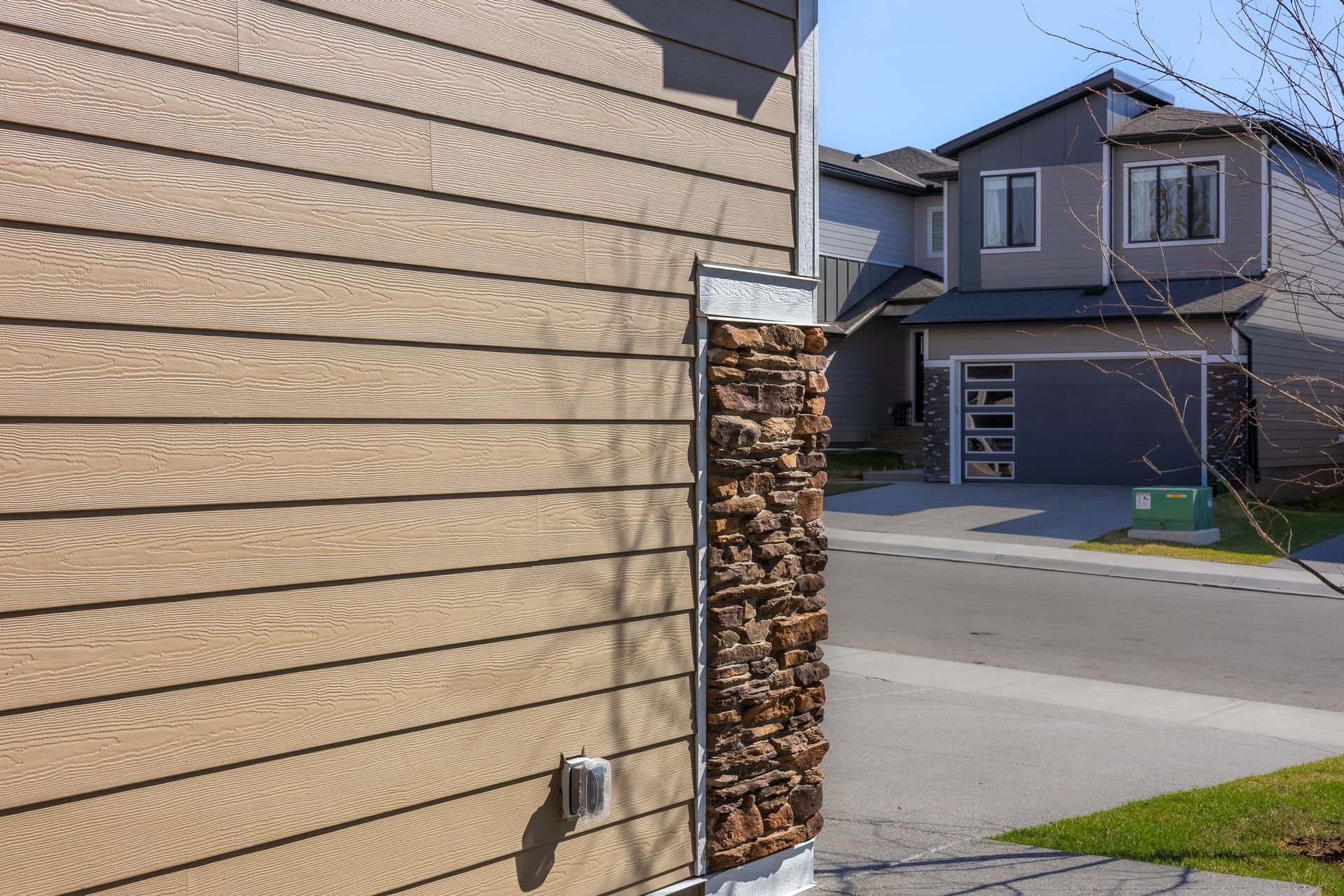House exterior with tan siding, stacked firewood by the wall, and a suburban street with neighboring homes House exterior with tan siding, stacked firewood by the wall, and a suburban street with neighboring homes