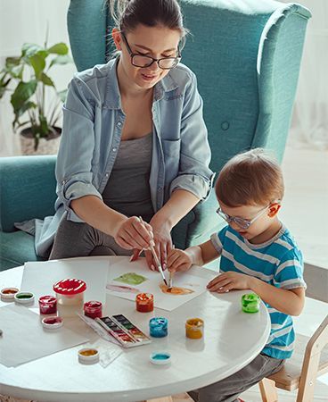 Woman and child painting at a table, both holding paintbrushes. Paints are in small pots on the table.