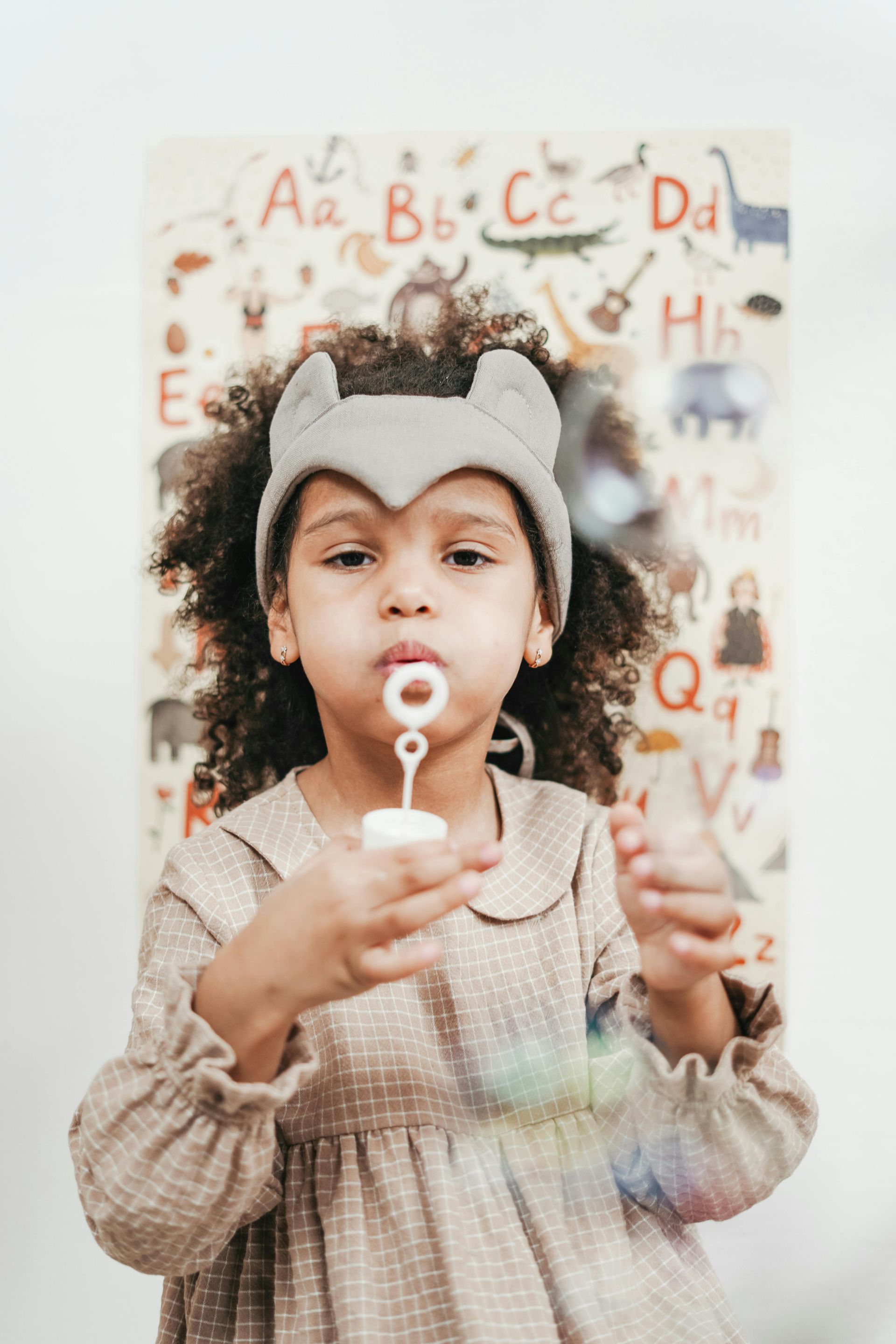 Child blowing bubbles in front of an alphabet poster, wearing a grey headband and a brown dress.