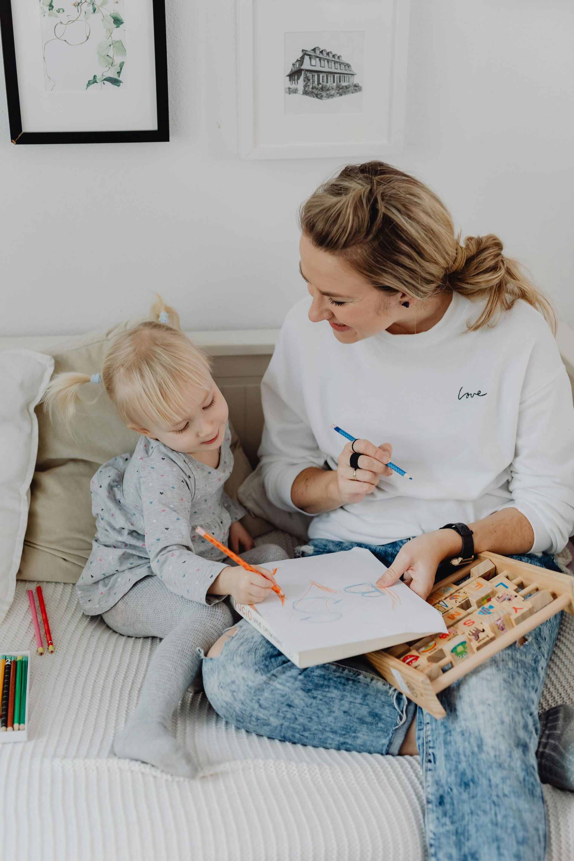 Woman and child drawing together on a couch, smiling, holding pens and looking at a notebook.