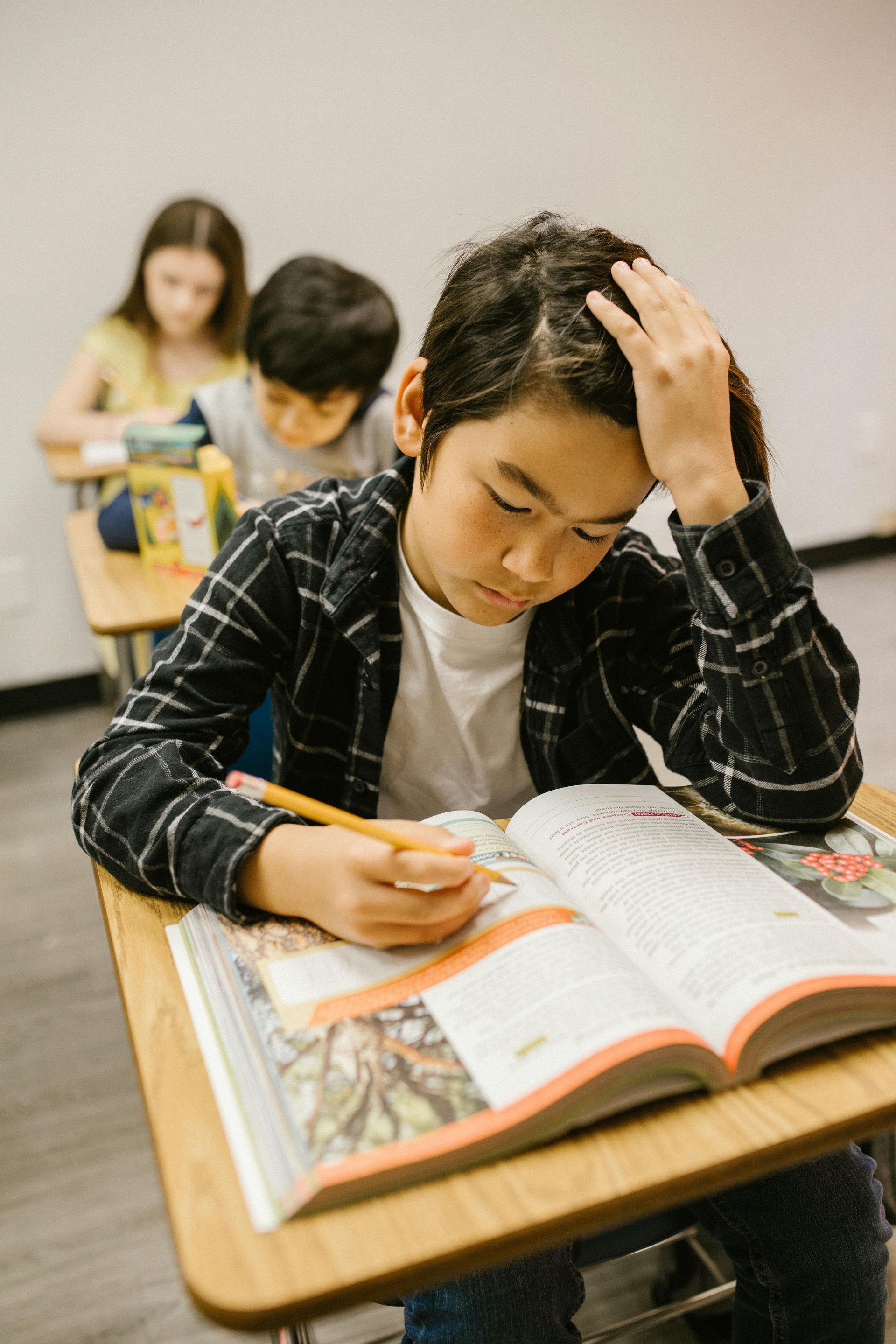 Student with hand on head, reading textbook in a classroom, other students in the background.