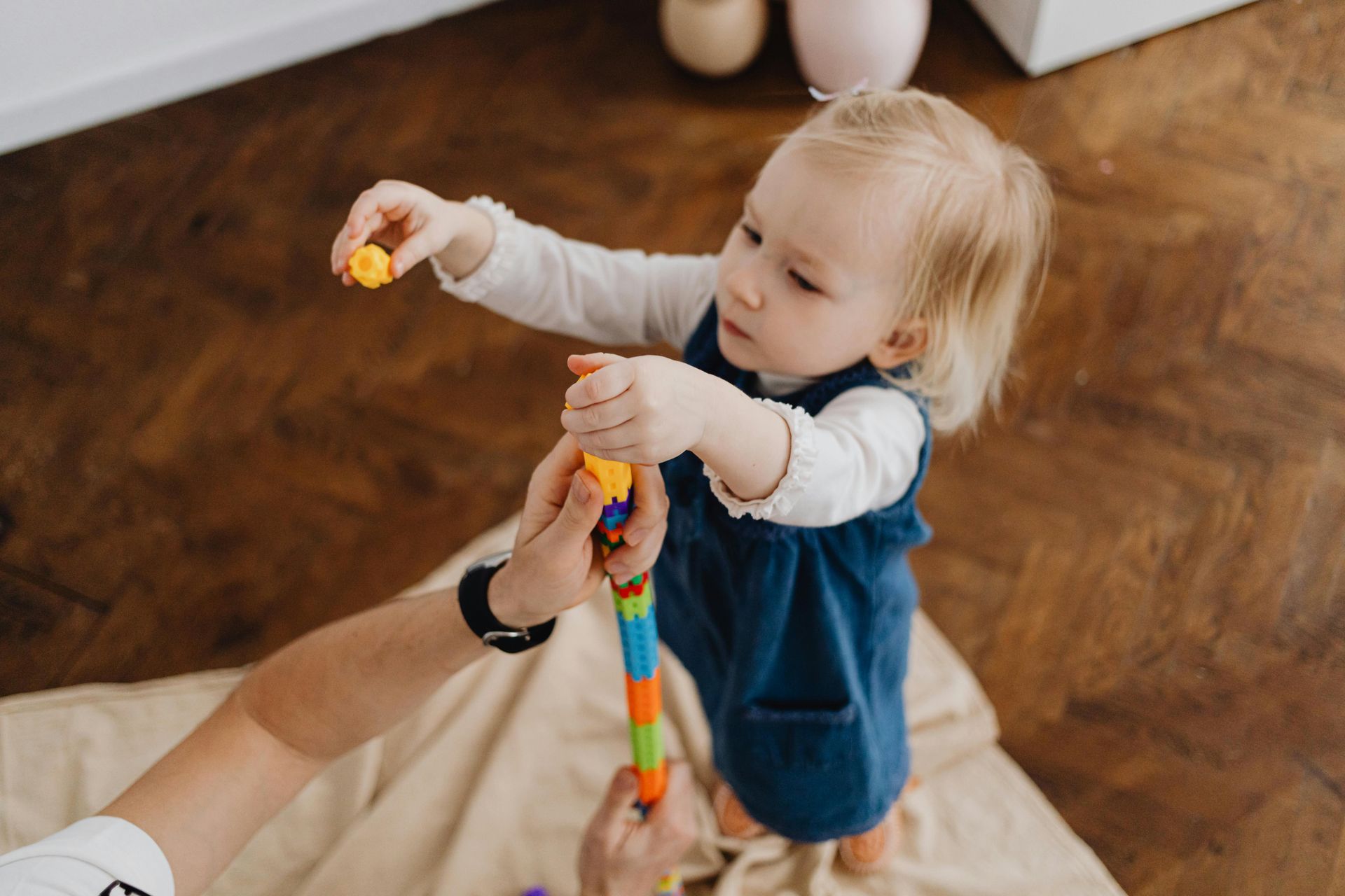 Child in blue jumper, playing with a colorful toy, held by an adult indoors on a wooden floor.