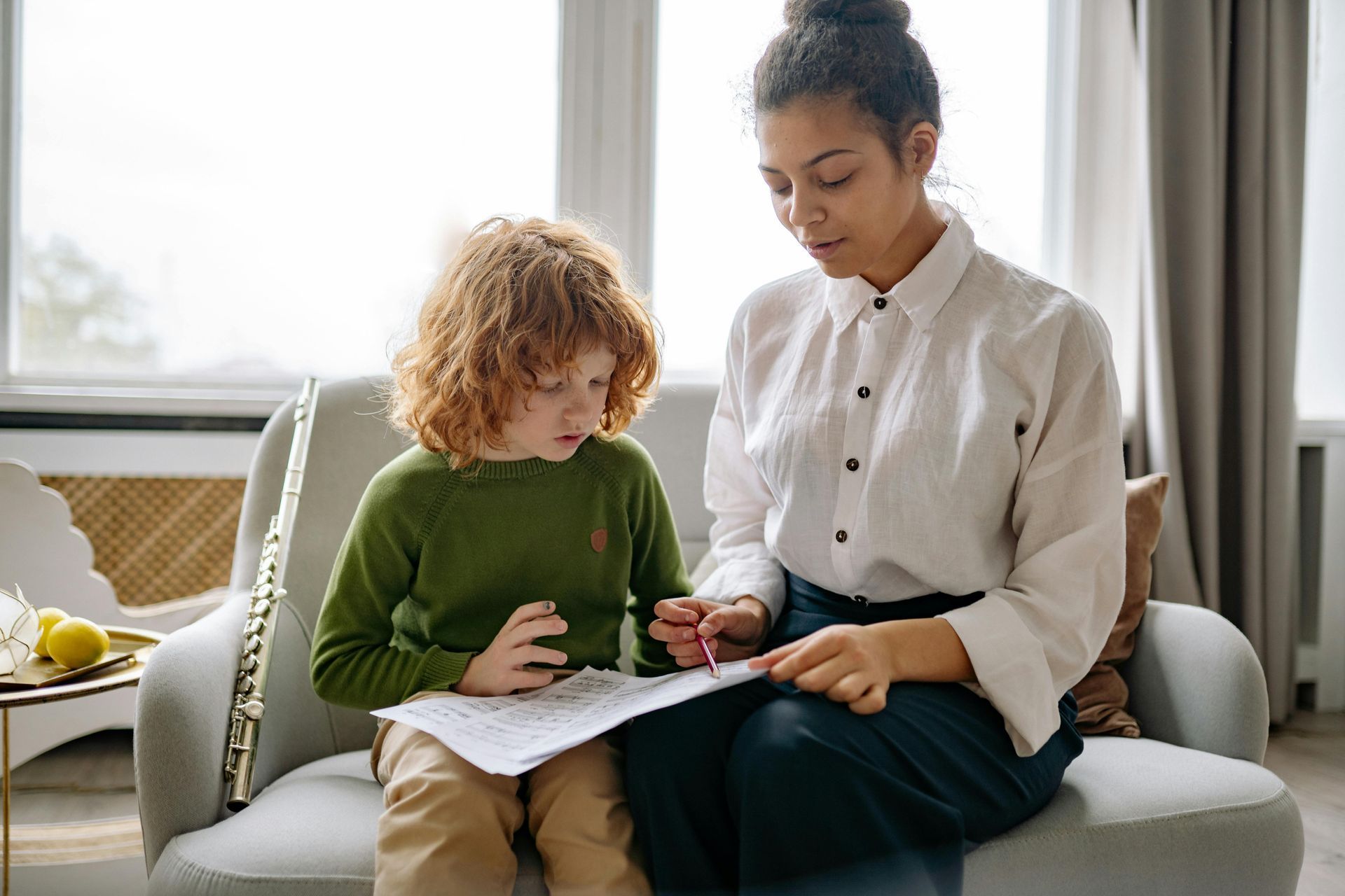 A person assists a child with music notes. They are seated on a couch near a window.