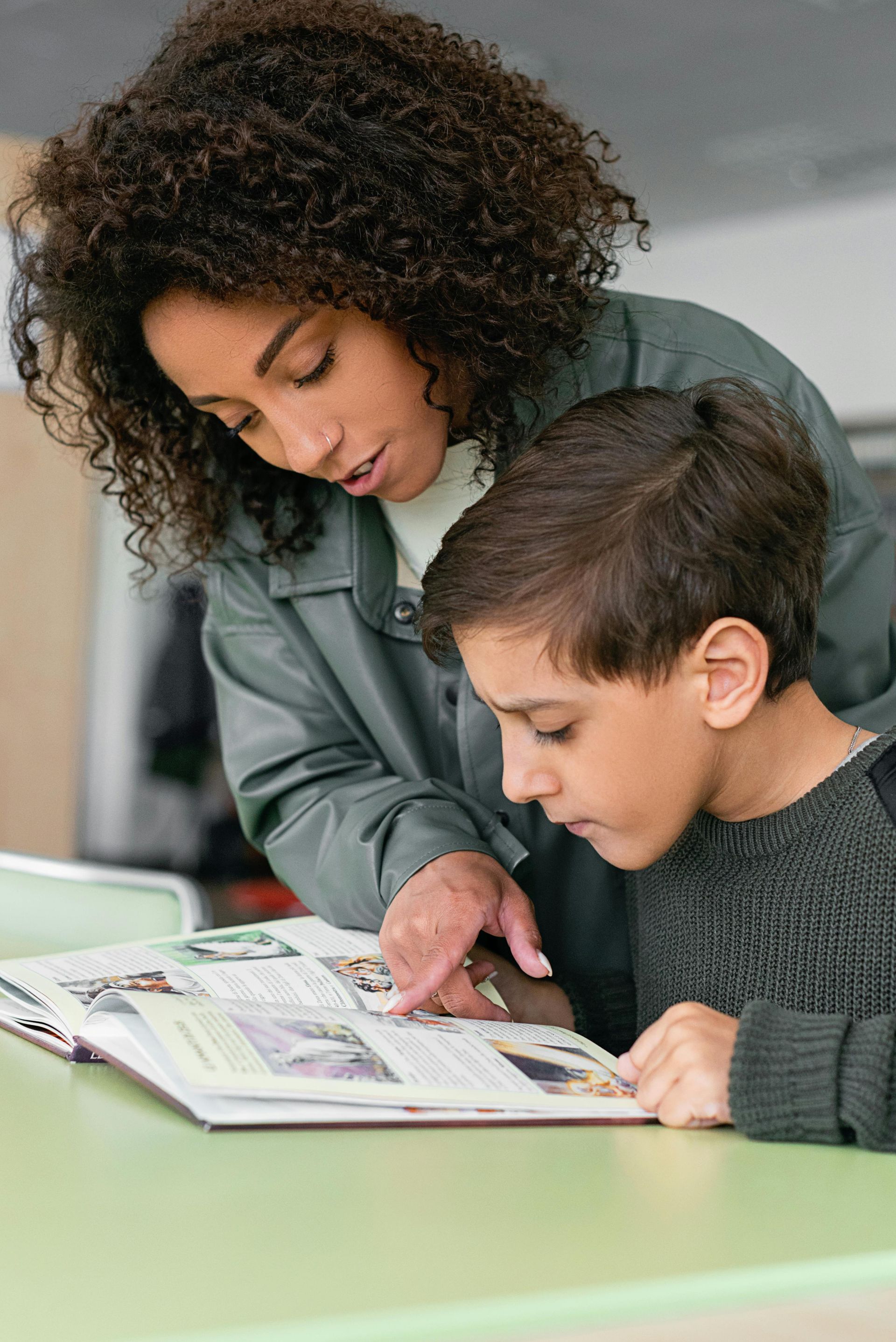 Woman points at book with boy, both looking at it attentively. Inside a room, light green table.
