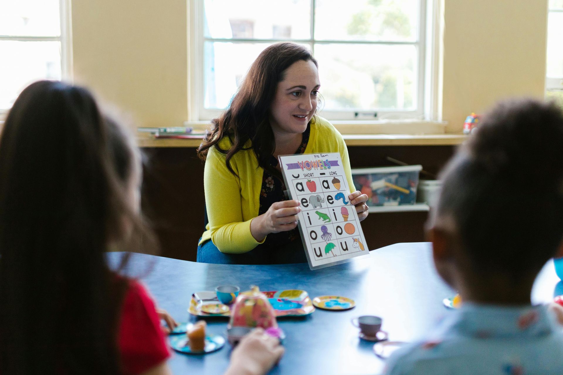 Teacher showing a poster of letters to children seated at a blue table. They are in a classroom.