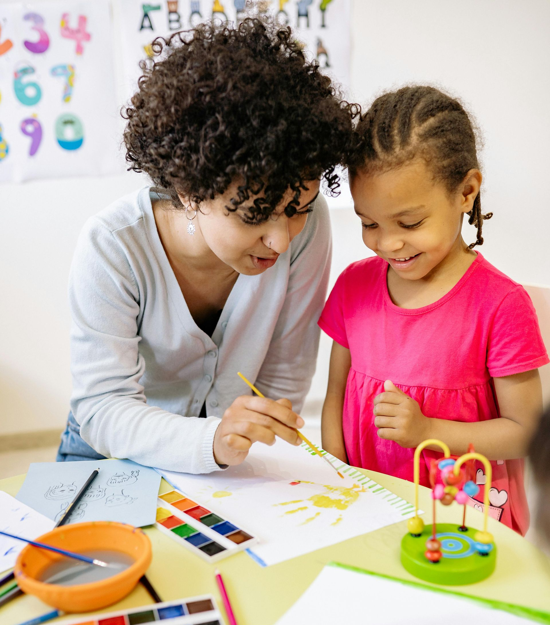 Woman helps child paint with watercolors at a table in a classroom.